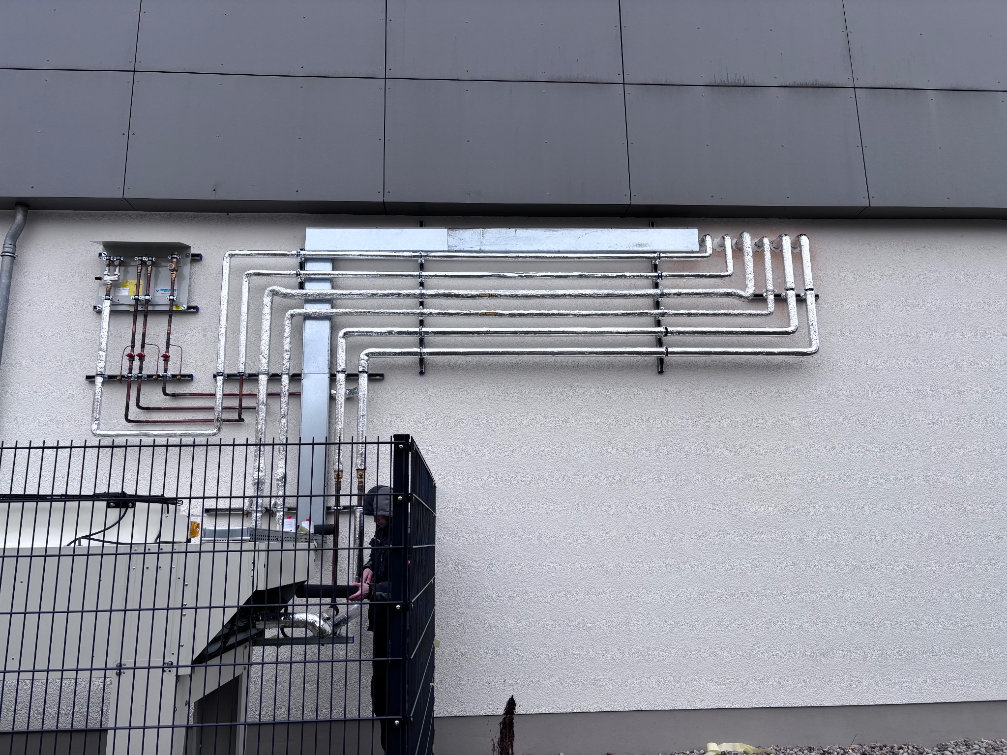 Zewnętrzna instalacja chłodnicza z rurami w srebrnej izolacji, zamontowana na jasnej ścianie budynku. Widoczny fragment urządzenia i osoba w ciemnym ubraniu za ogrodzeniem.