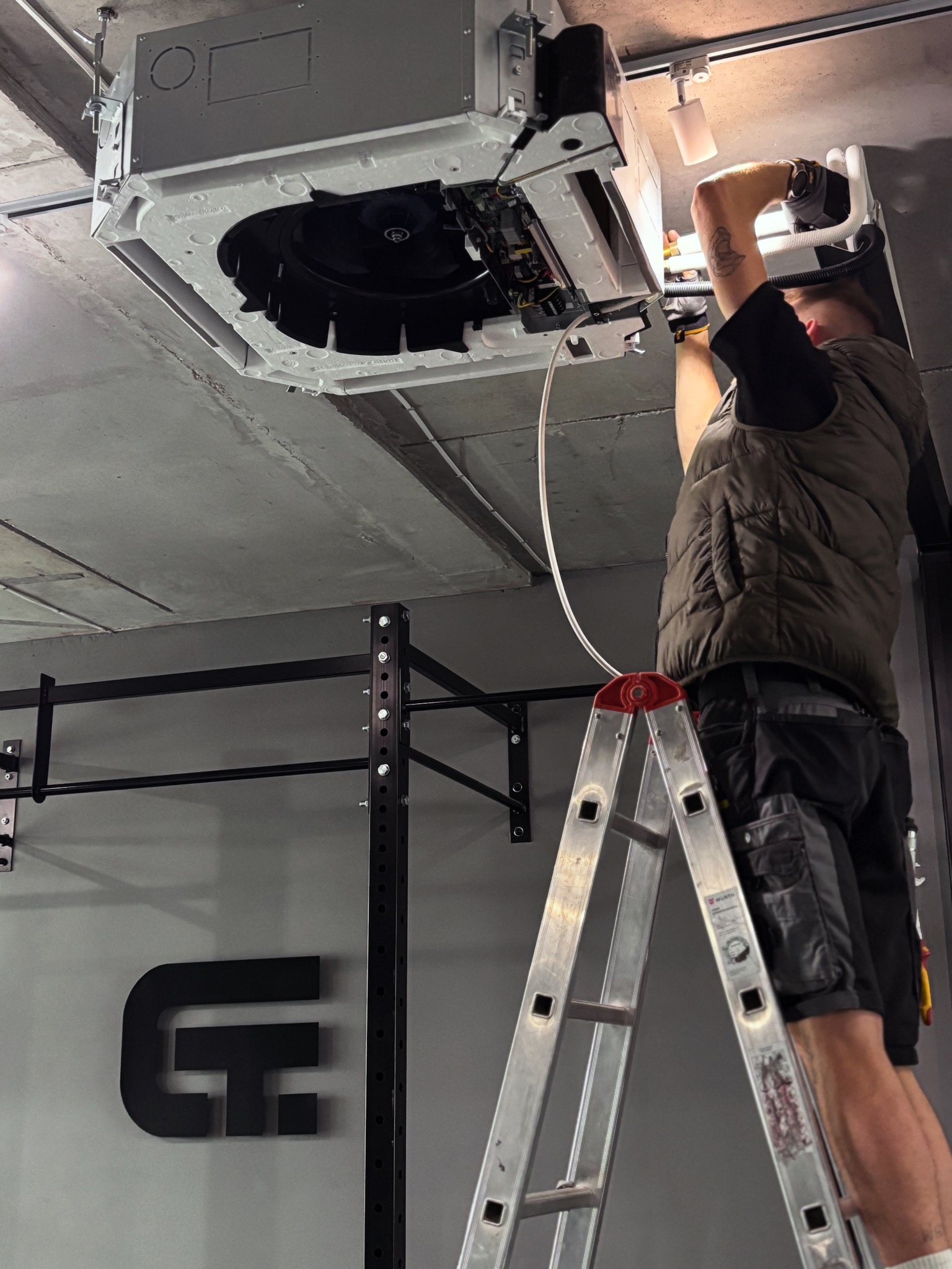 Montaż sufitowej klimatyzacji kasetonowej w szarej przestrzeni, widoczny pracownik na drabinie i fragment konstrukcji do ćwiczeń. Oświetlenie punktowe.