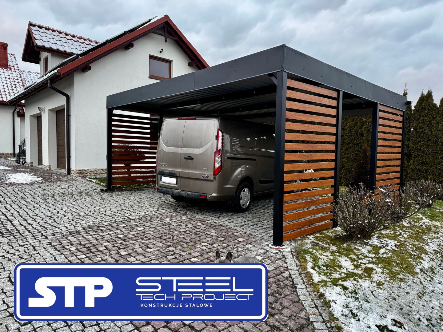 Nowoczesna, czarno-drewniana konstrukcja z metalu, pełniąca funkcję zadaszenia dla samochodu, na tle domu z czerwoną dachówką i kostki brukowej.