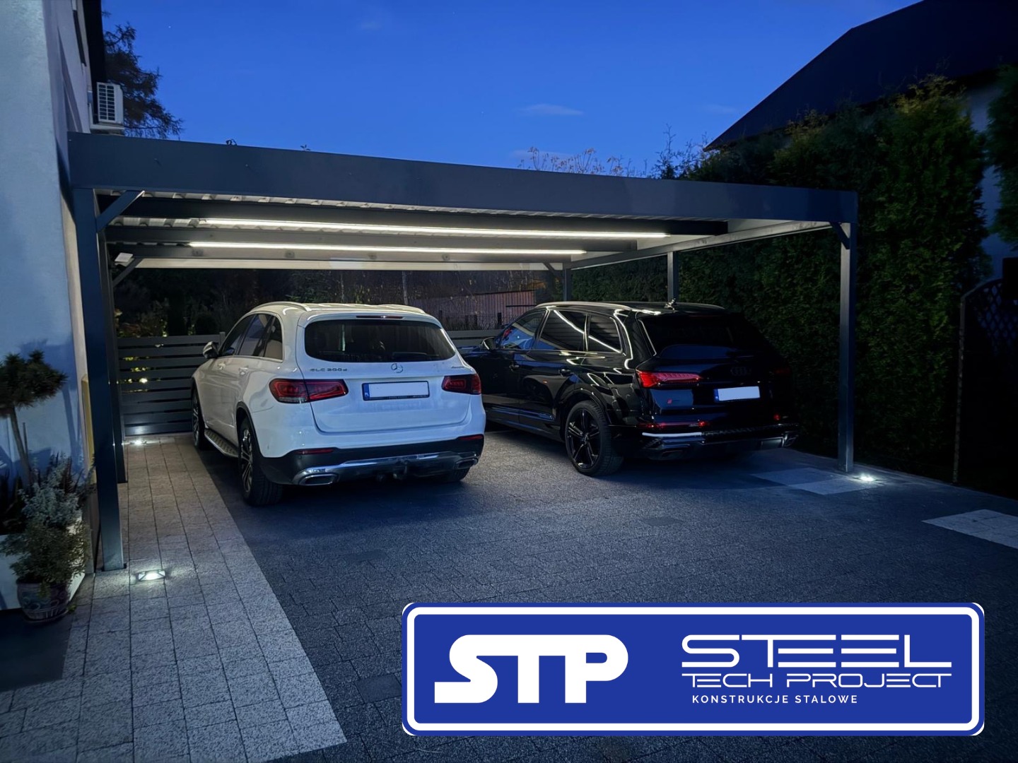 Nowoczesna, grafitowa konstrukcja z oświetleniem LED, osłaniająca dwa samochody: biały Mercedes i czarne Audi, na posesji z kostki brukowej.