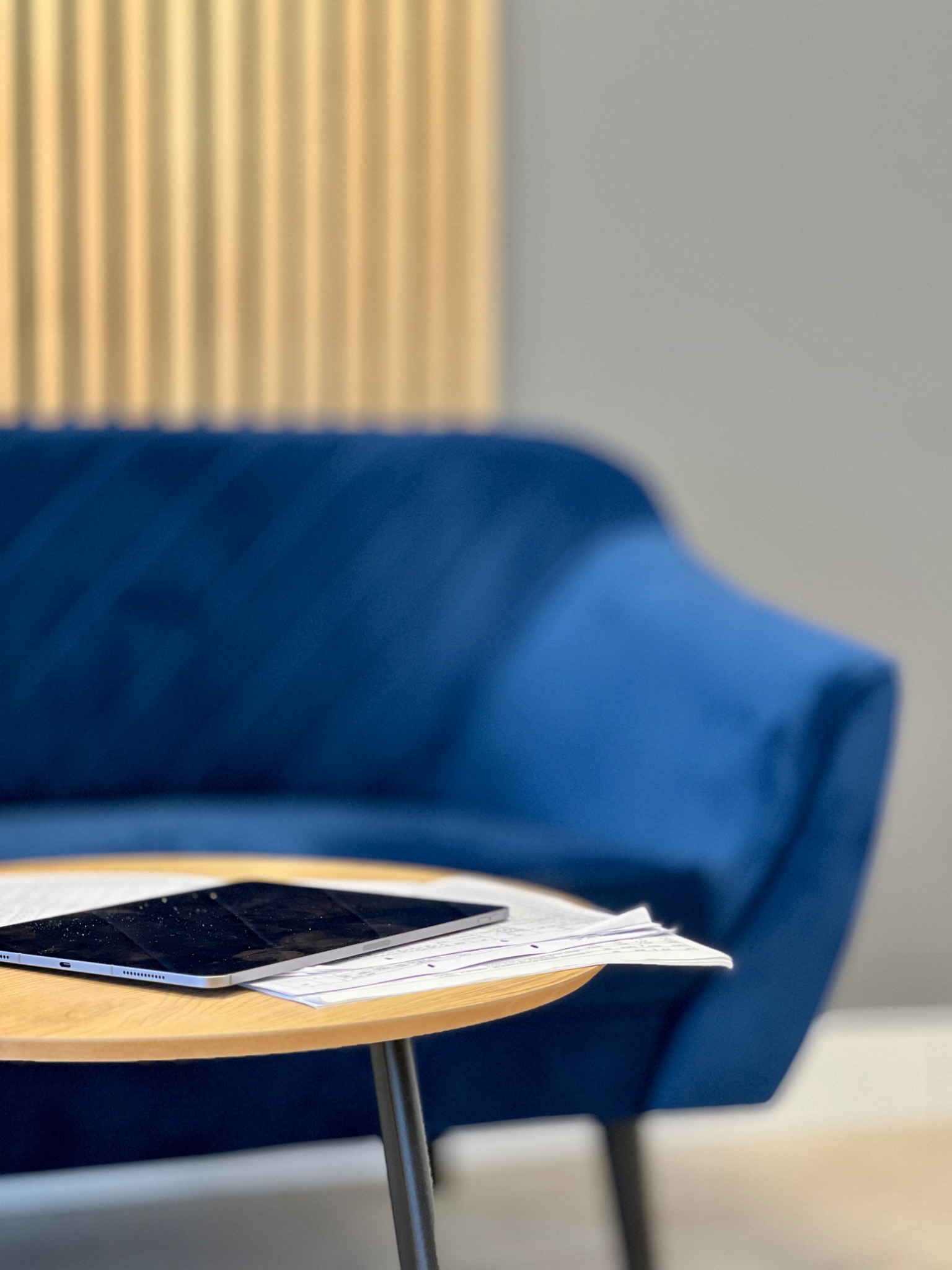Tablet i dokumenty na okrągłym stoliku kawowym, w tle niebieska sofa i dekoracyjna ściana. Nowoczesne biuro, gotowość do pracy.