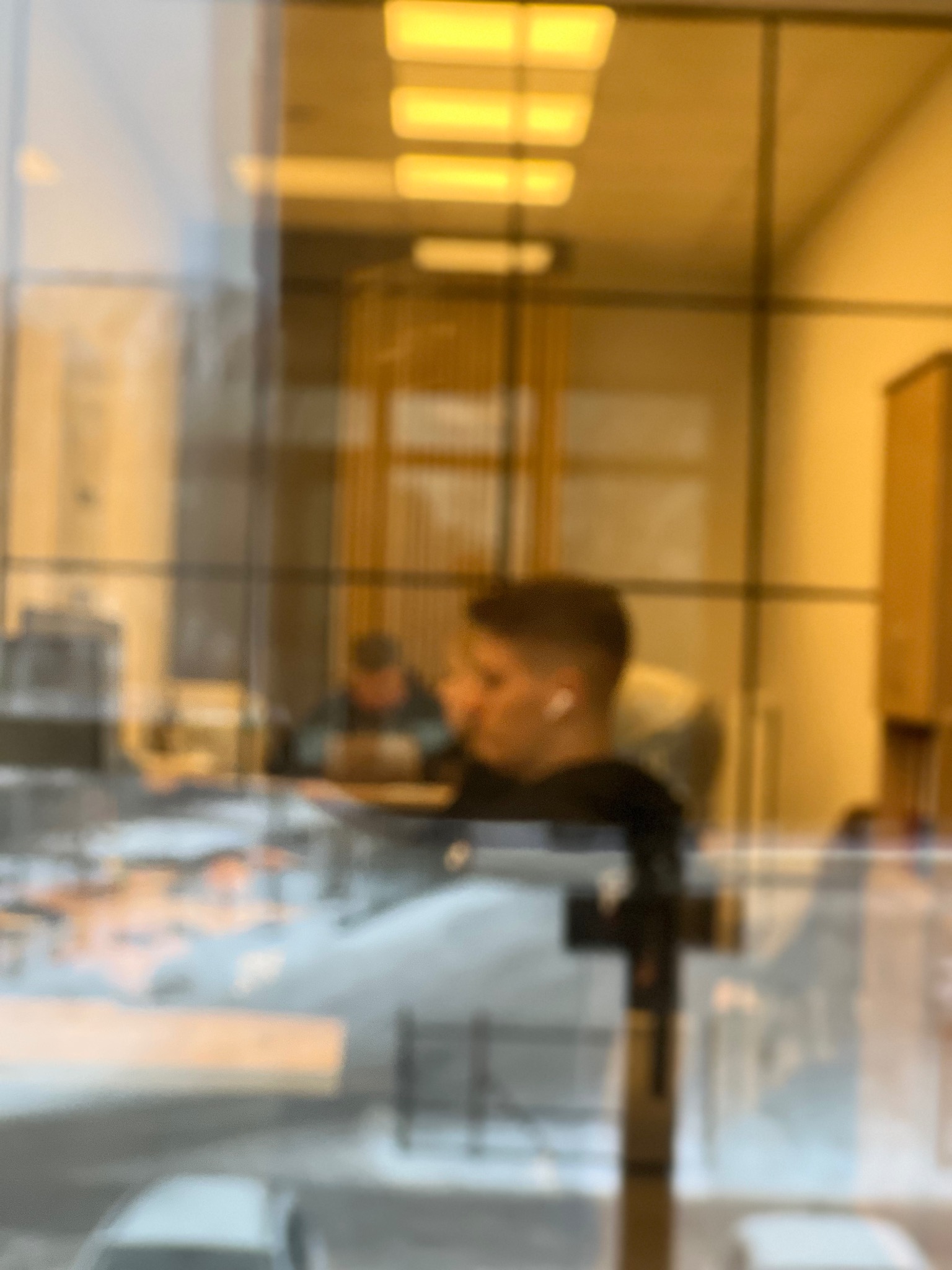 Rozmyte odbicie biura przez szybę: pracownicy przy biurkach, ciepłe oświetlenie, nowoczesny wystrój. Widok z zewnątrz, lekka abstrakcja.