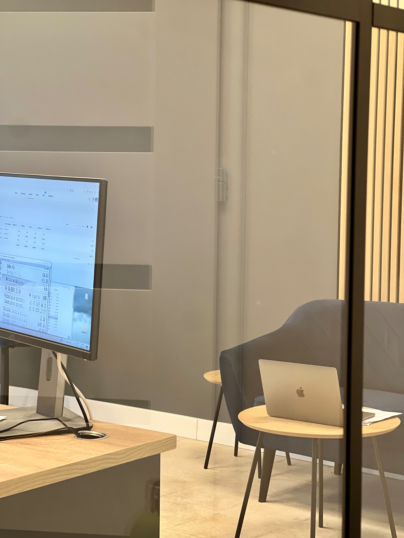 Nowoczesne biuro rachunkowe: stanowisko pracy z komputerem wyświetlającym dane finansowe, laptop na stoliku i sofa w tle, oddzielone szklaną ścianką.