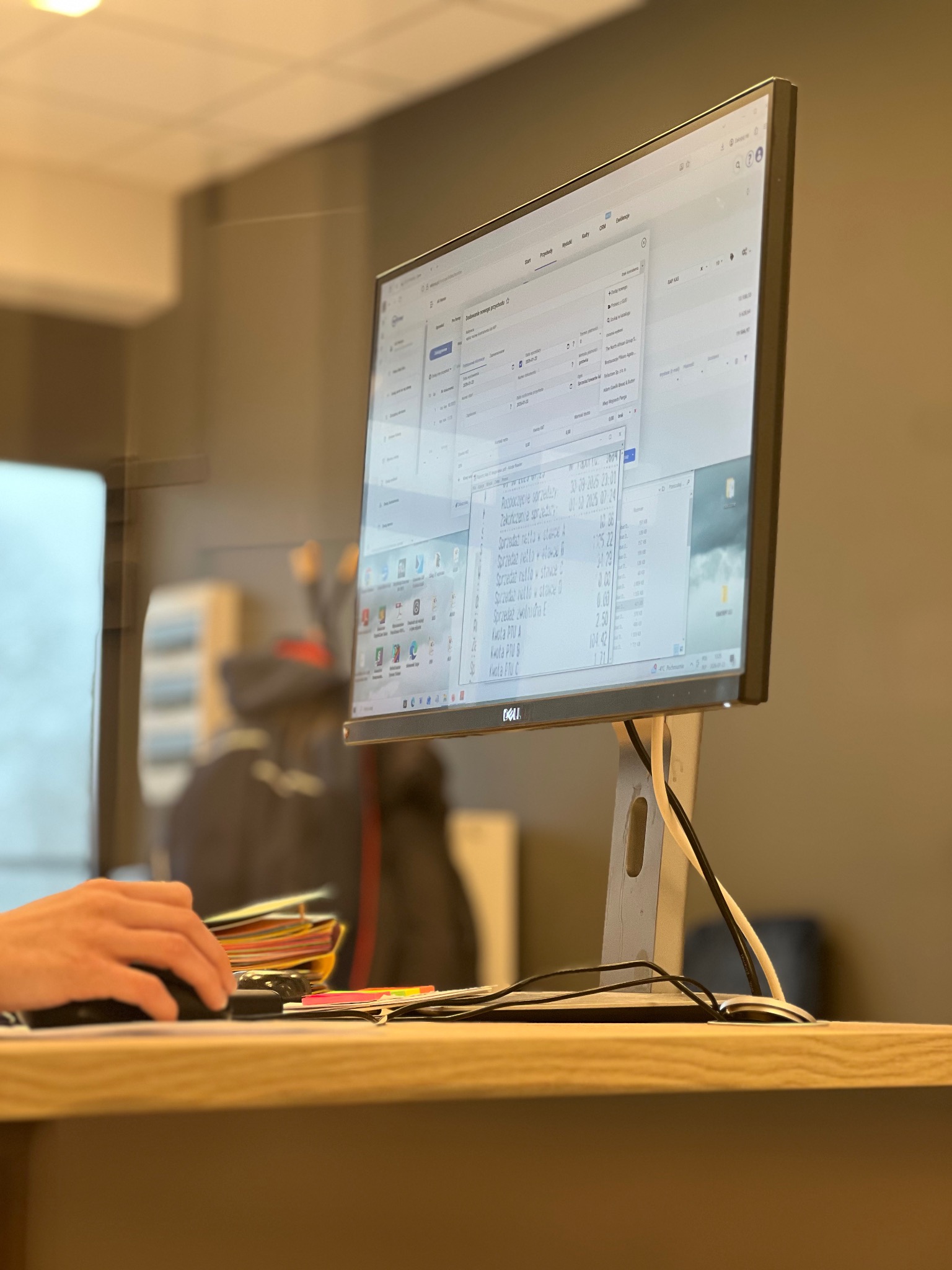 Praca biurowa: dłoń obsługuje mysz przed monitorem Dell, na którym widoczny jest program księgowy z danymi sprzedaży. Obok stos dokumentów i notatki.