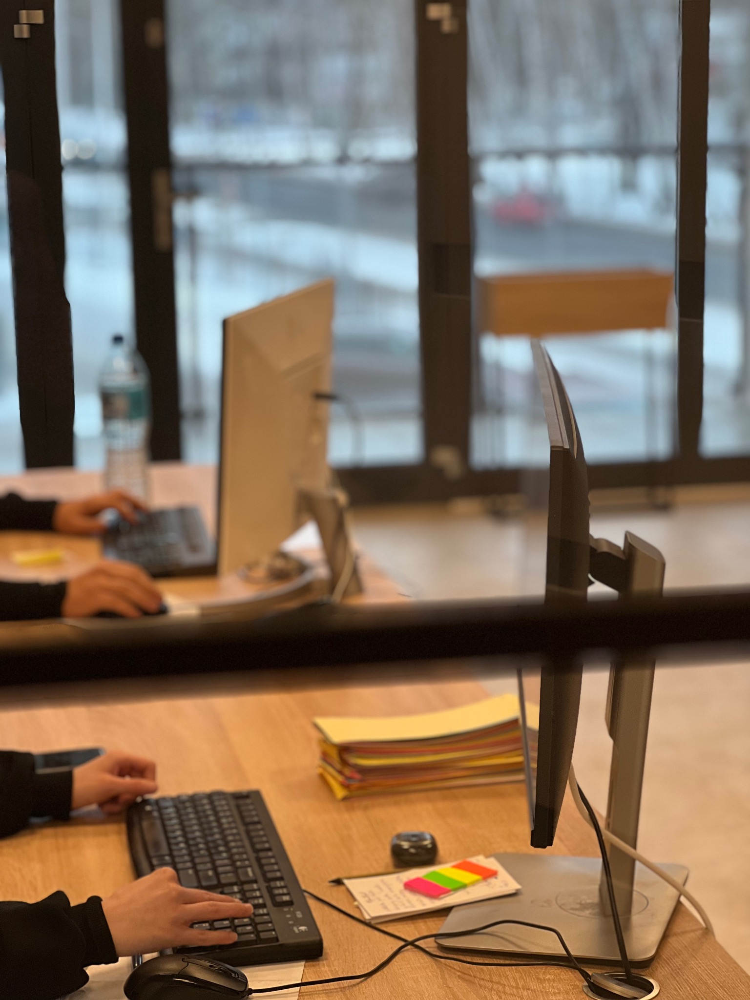 Biuro z pracownikami przy komputerach, stos dokumentów, notatki samoprzylepne. Praca w skupieniu, jasne wnętrze z widokiem na zewnątrz.