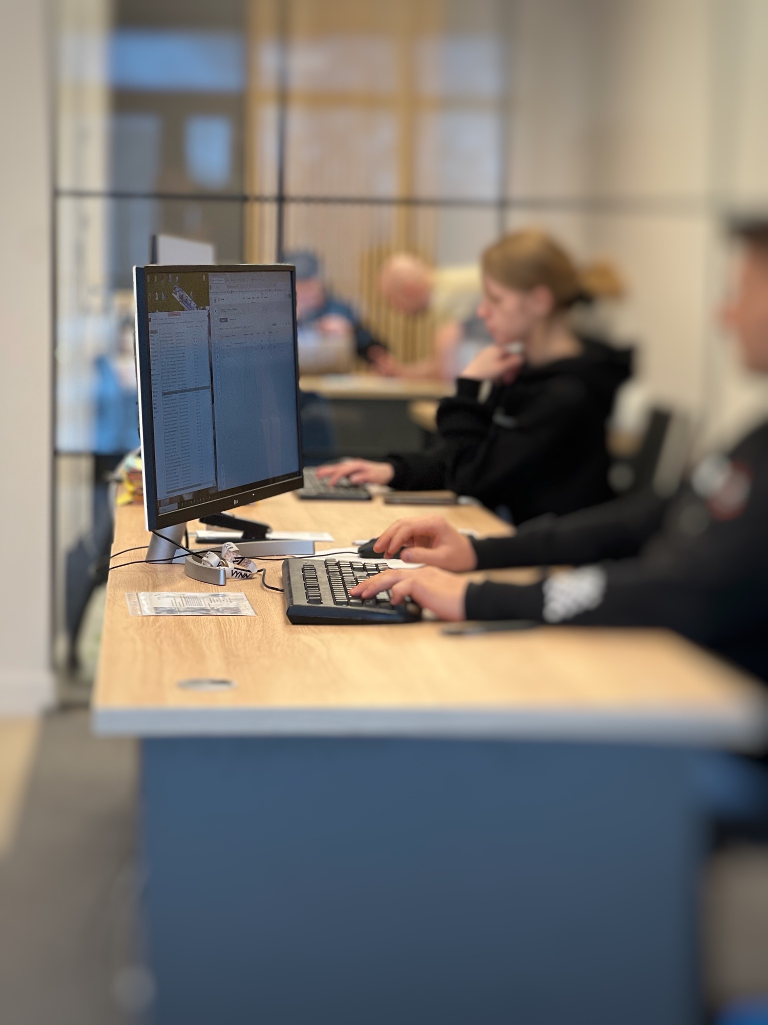 Praca biurowa: osoba przy komputerze, dłonie na klawiaturze, monitor z danymi, w tle inni pracownicy. Jasne biuro, nowoczesny design.