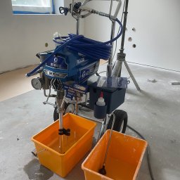 ATŁABUD - Agregat malarski Graco ST MAX II 595 PC Pro z niebieskim wężem i dwoma żółtymi pojemnikami na farbę w pomieszczeniu w trakcie malowania. Widoczne ślady farby.