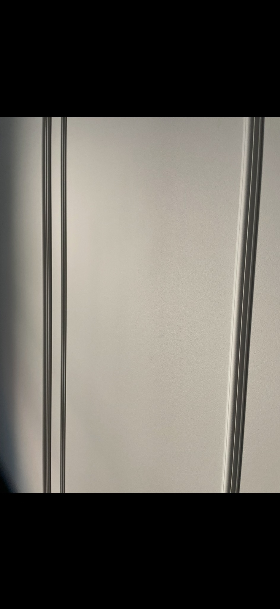 Fragment białej ściany z delikatną fakturą i pionowymi, srebrnymi listwami dekoracyjnymi po bokach. Minimalistyczny detal wykończenia wnętrza.