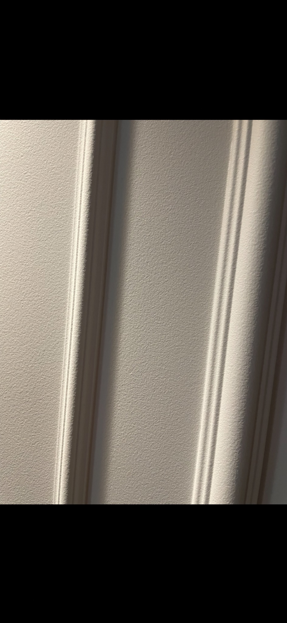 Fragment białej ściany z dekoracyjnymi listwami w pionowym ujęciu, oświetlony bocznym światłem, tworzącym wyraźne cienie.