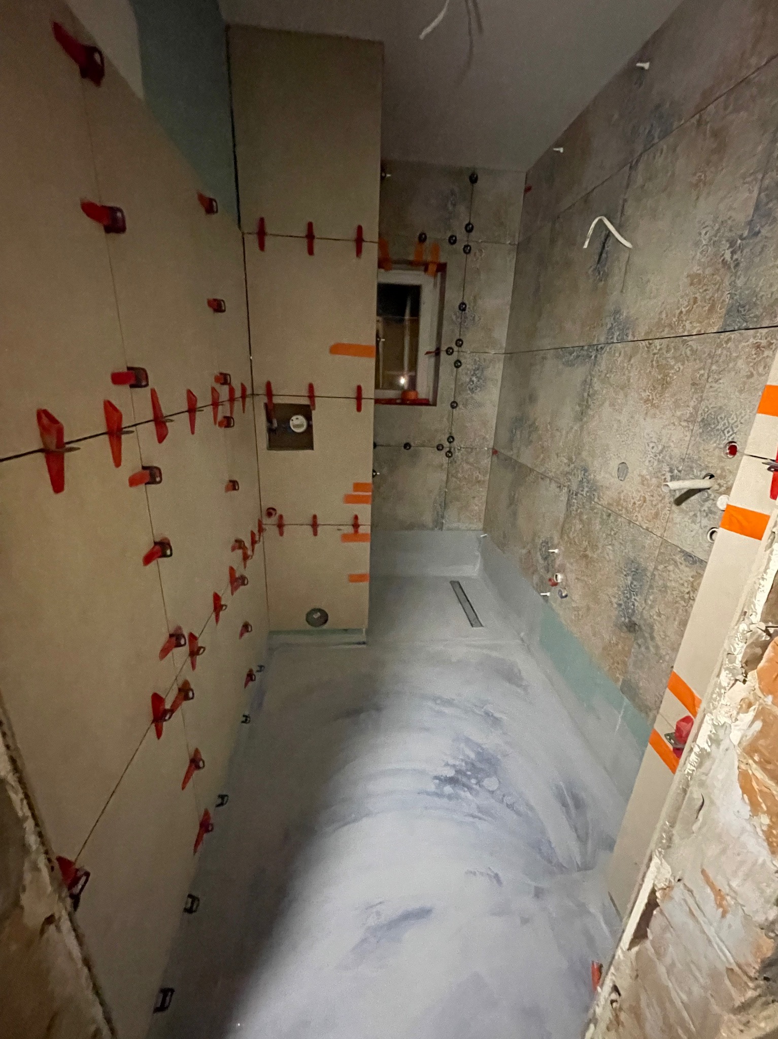 Wnętrze łazienki w trakcie prac glazurniczych. Widoczne płytki z użyciem systemu poziomowania, odpływ liniowy w posadzce i fragment okna.