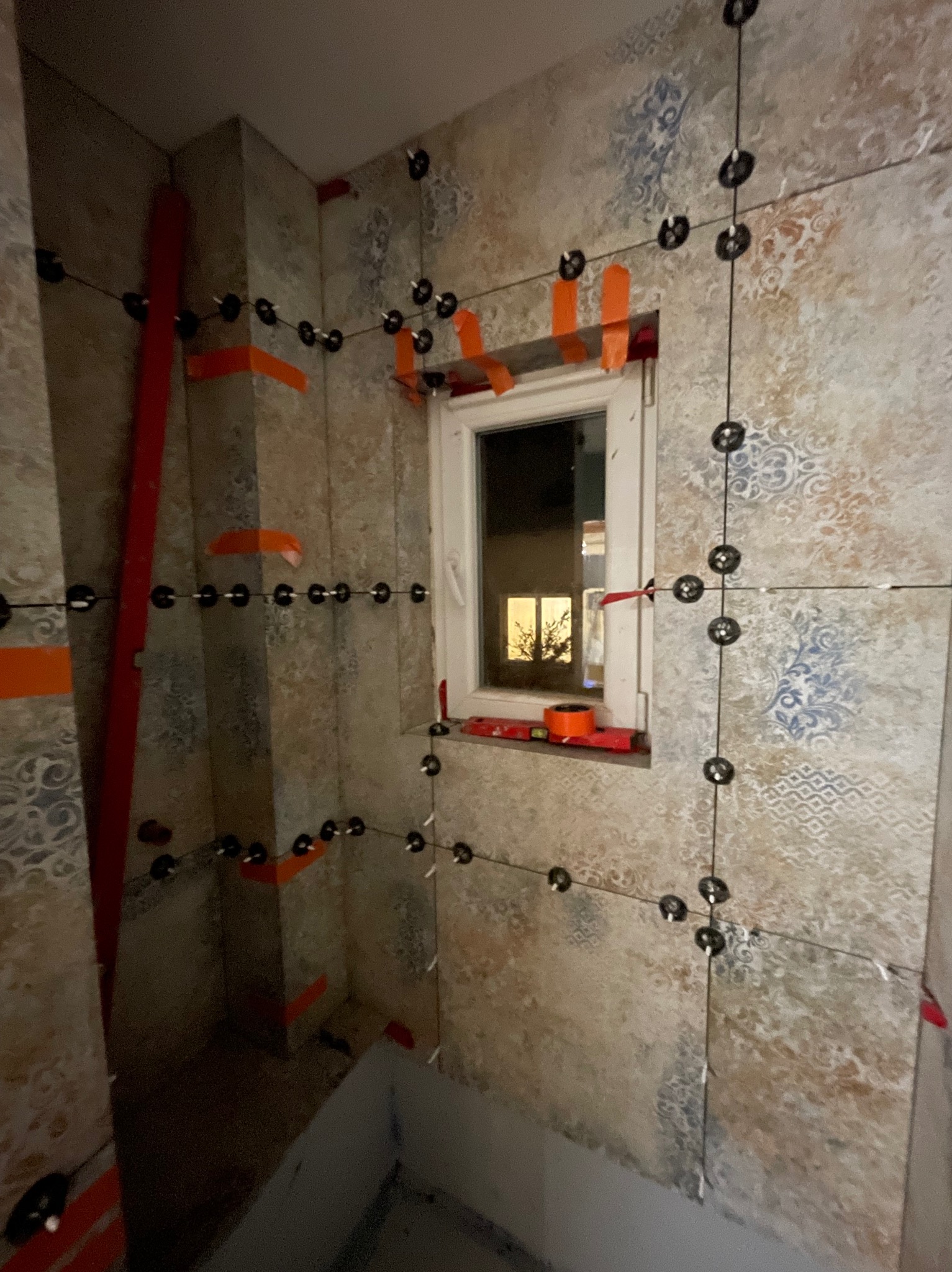 Ściana łazienki w trakcie układania płytek z wzorem, z użyciem systemu poziomowania. Widoczne czarne klipsy i pomarańczowe paski mocujące przy oknie.