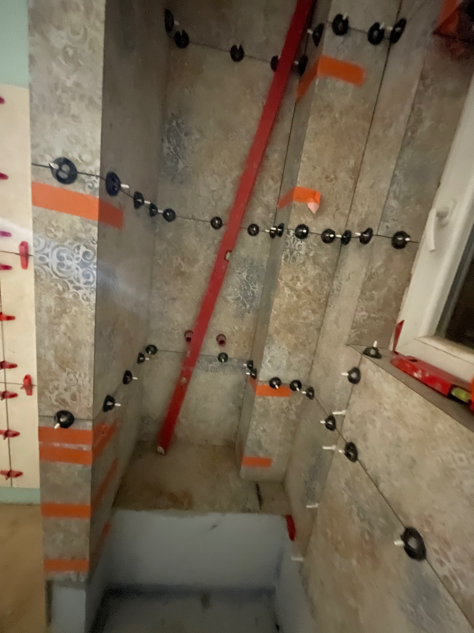 Układanie płytek w narożniku wnęki okiennej, z użyciem poziomicy i systemu poziomowania płytek. Widoczne pomarańczowe paski i czarne klipsy.