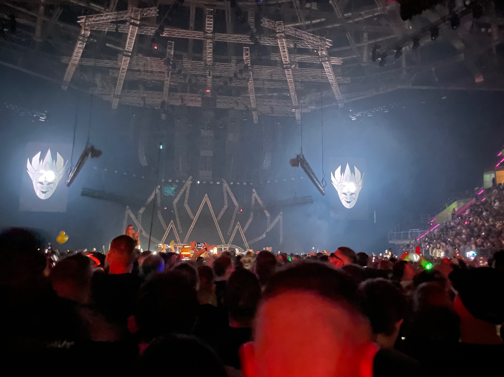 Widok na scenę z tłumu podczas koncertu. W tle dekoracje sceniczne i oświetlenie. Widać publiczność z podniesionymi rękami i telefonami.