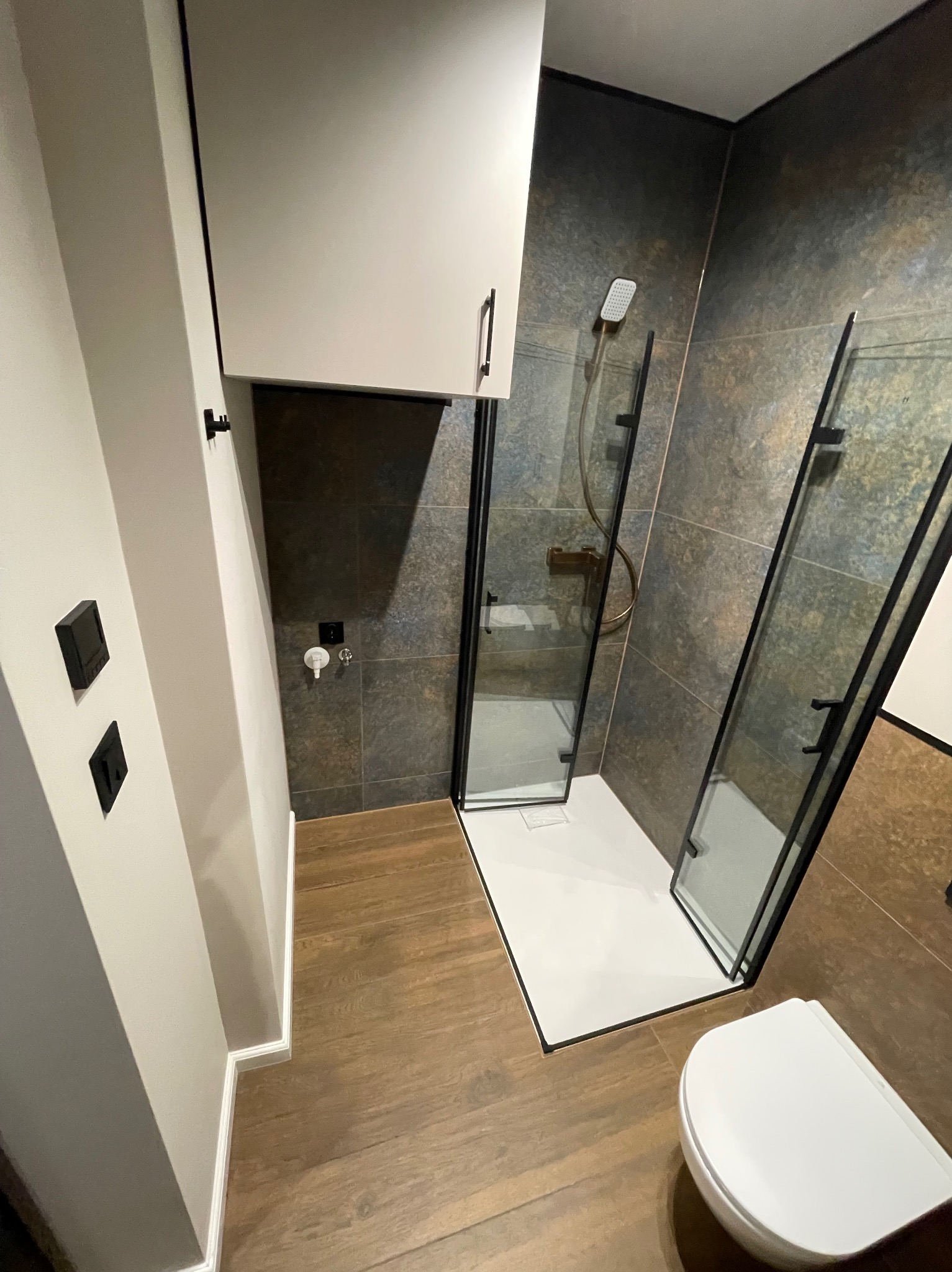 Nowoczesna łazienka z kabiną prysznicową typu walk-in, wykończona płytkami w odcieniach brązu i beżu. Widoczna biała umywalka i drewnopodobna podłoga.