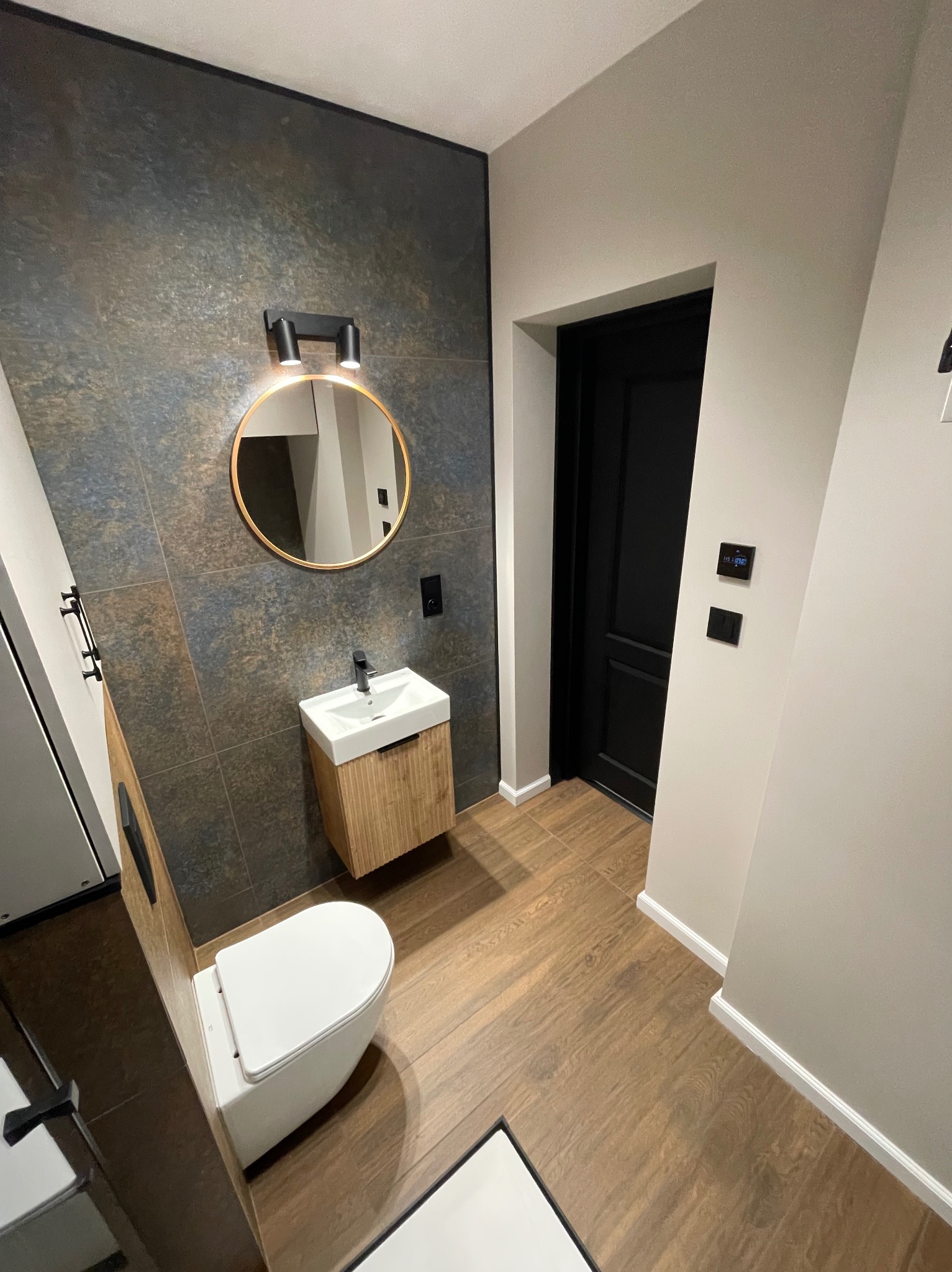 Nowoczesna łazienka z płytkami imitującymi kamień, okrągłym lustrem z podświetleniem i drewnianą szafką pod umywalką. Ciemne drzwi i drewnopodobna podłoga.