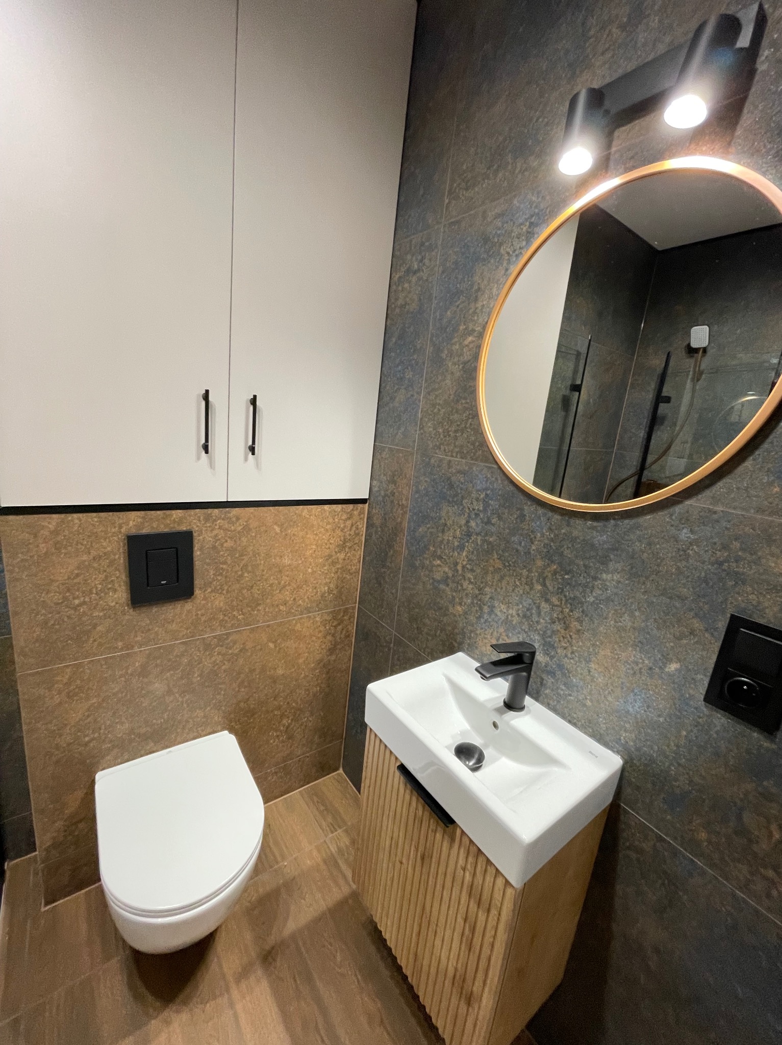 Nowoczesna łazienka po remoncie: wisząca toaleta, umywalka z szafką z lamelami, okrągłe lustro z podświetleniem i płytki imitujące kamień w odcieniach brązu i szarości.