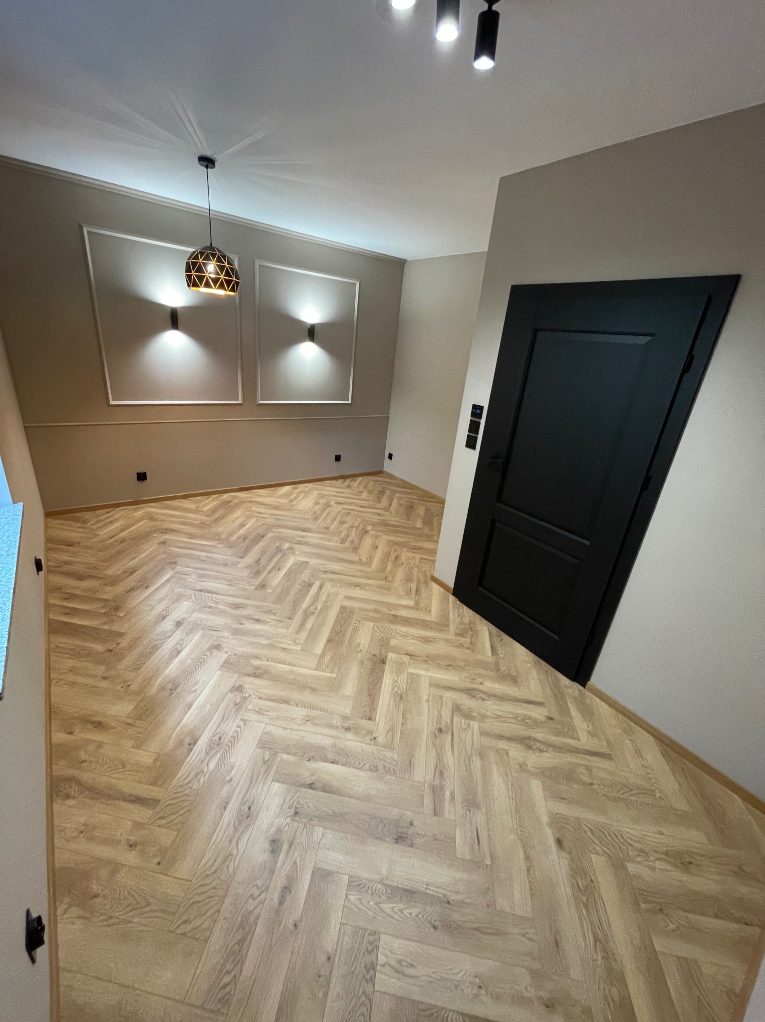 Jasne panele ułożone w jodełkę, czarne drzwi zlicowane ze ścianą, oświetlenie sufitowe i dekoracyjne listwy ścienne w wykończonym wnętrzu.