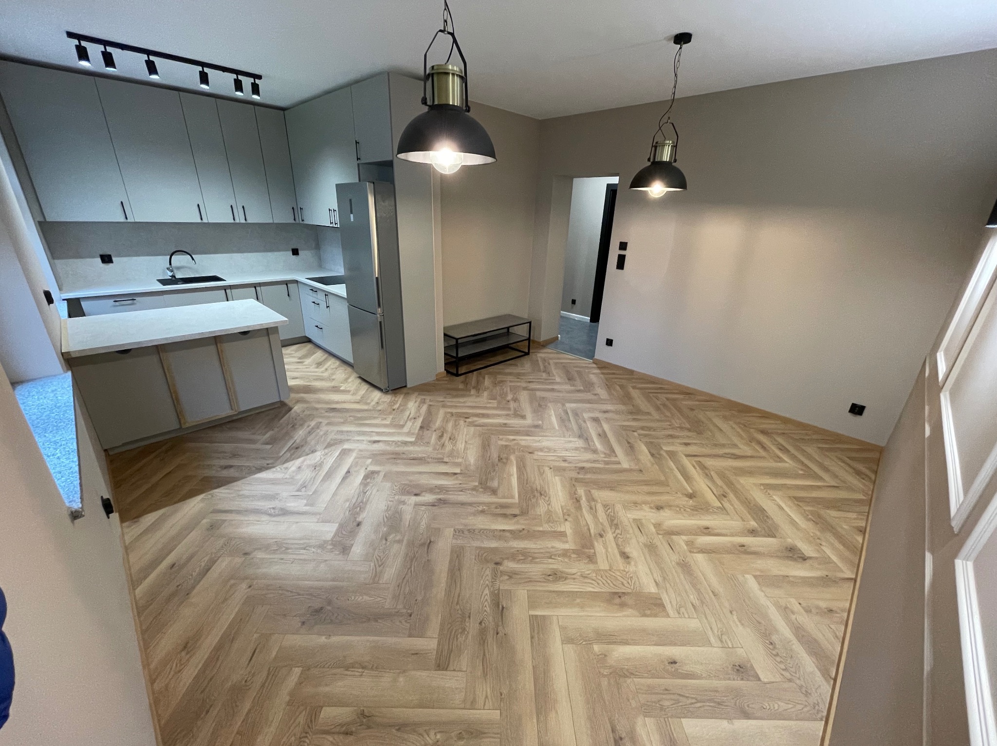 Jasne panele ułożone we wzór jodełki francuskiej w przestronnym wnętrzu z szarą zabudową kuchenną i nowoczesnym oświetleniem. Widok na korytarz.