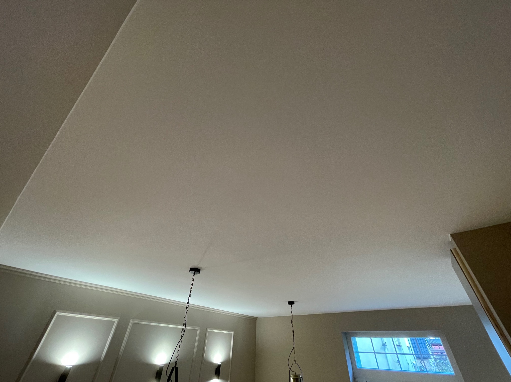 Gładka, jasna powierzchnia sufitu po szpachlowaniu, widoczne lampy wiszące i okno z widokiem na zewnątrz. Minimalistyczne wykończenie wnętrza.