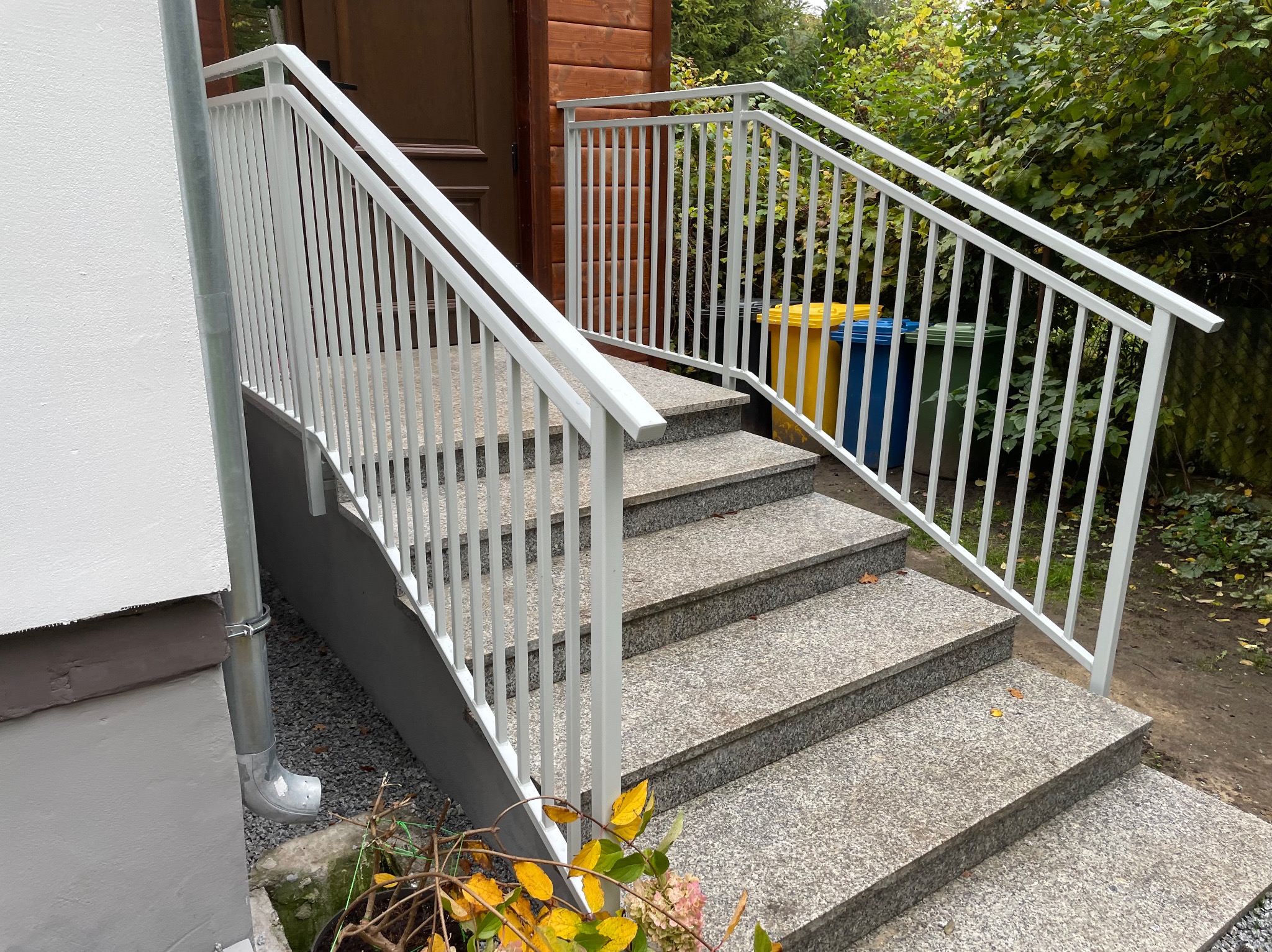 Granitowe schody zewnętrzne z białą, metalową balustradą, prowadzące do drewnianego budynku. Widoczne kosze na śmieci w tle i rynna przy ścianie.