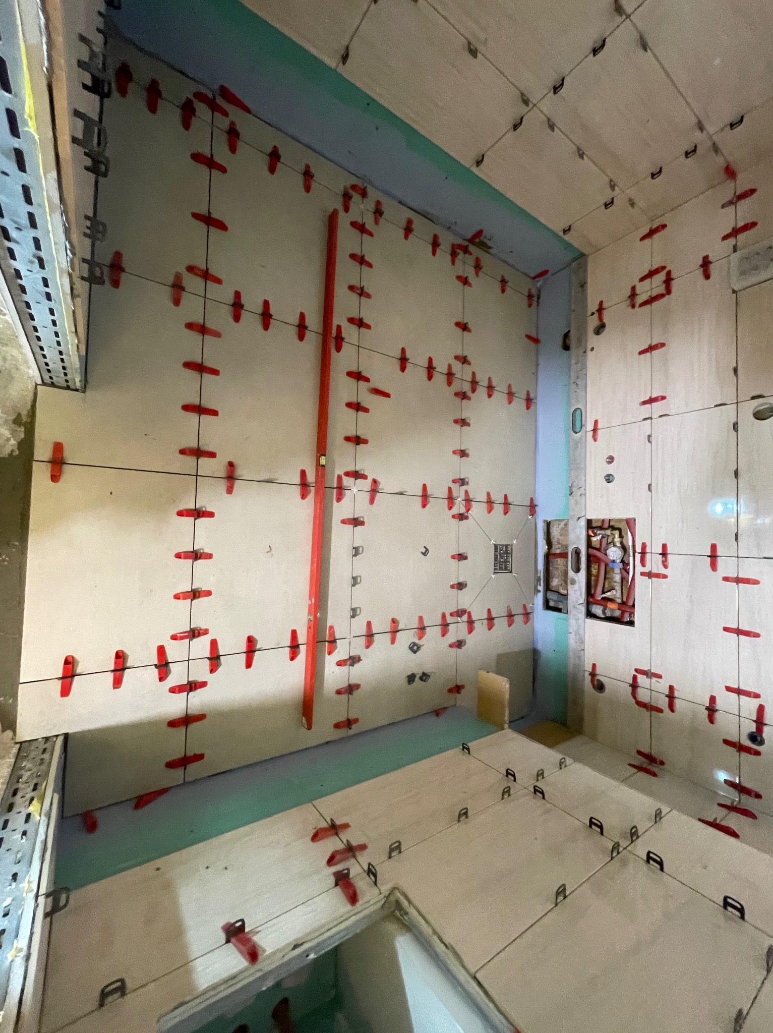 Świeżo ułożone, jasne płytki w łazience, z czerwonymi krzyżykami dystansowymi i systemem poziomowania. Widoczne piony i poziomy, instalacja wodna w ścianie.