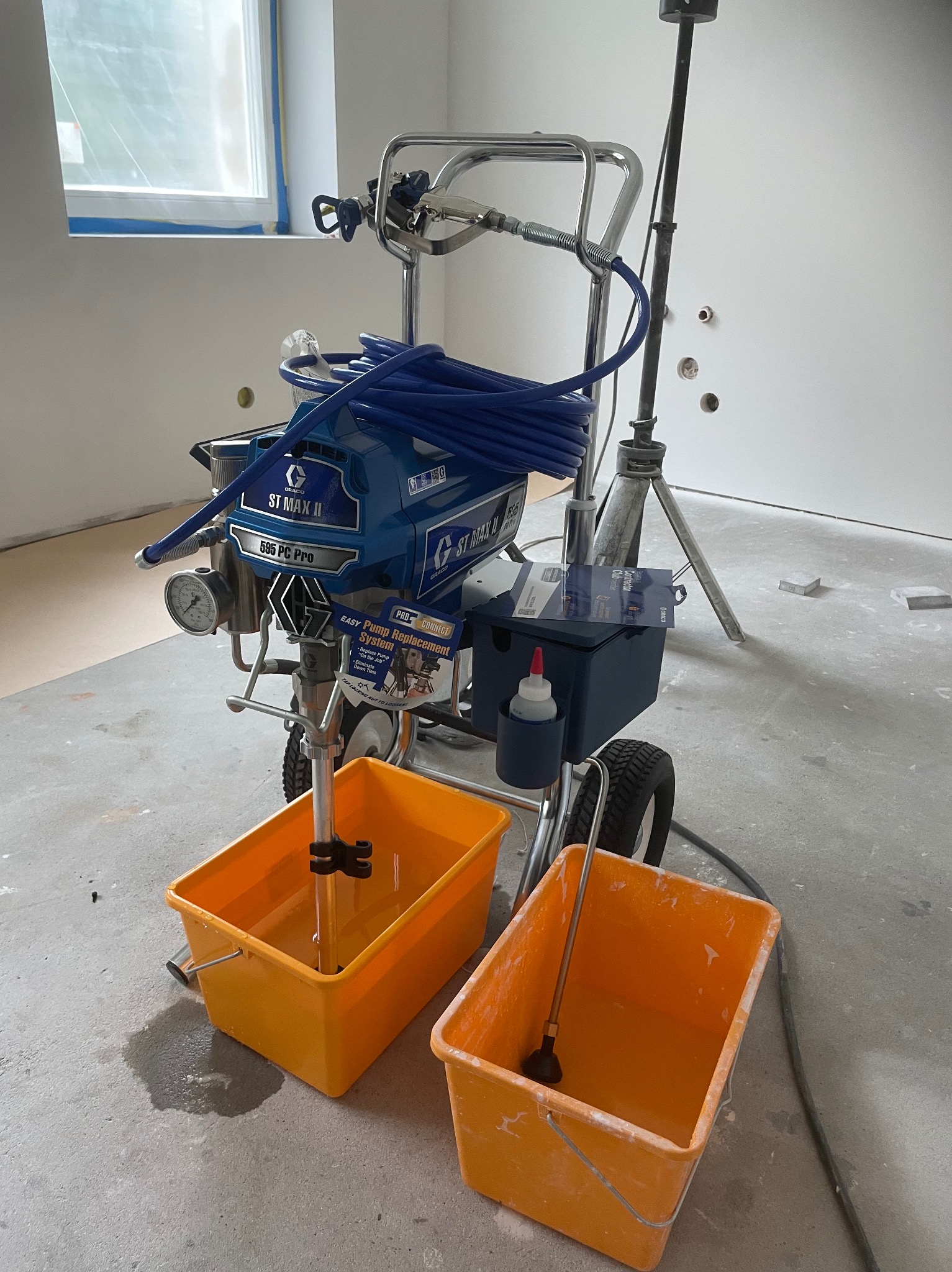 Agregat malarski Graco ST MAX II 595 PC Pro z niebieskim wężem i dwoma żółtymi pojemnikami na farbę w pomieszczeniu w trakcie malowania. Widoczne ślady farby.