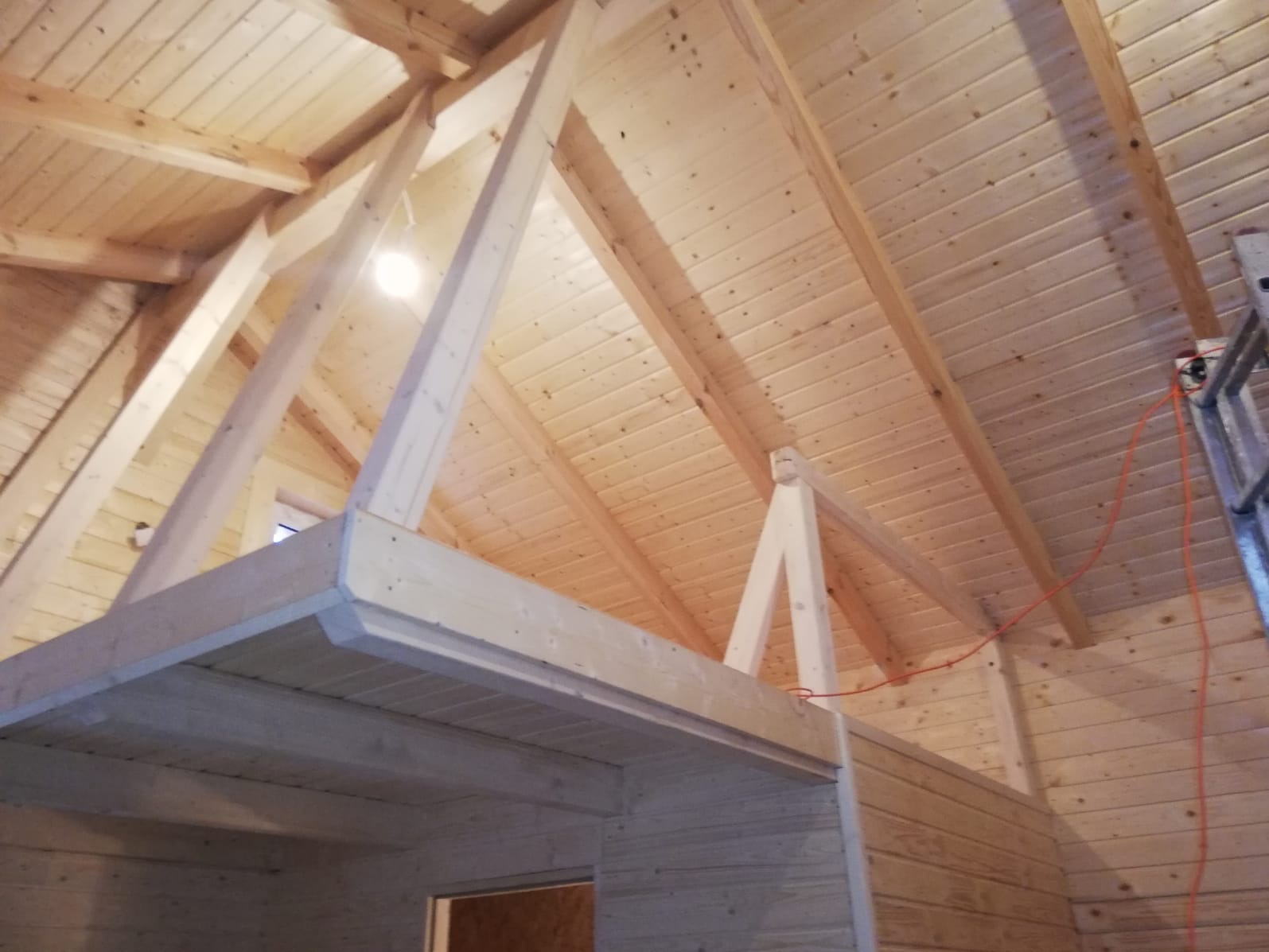 Drewniane wnętrze z widoczną konstrukcją dachu i antresolą, wykończone boazerią. Widoczne elementy instalacji elektrycznej i drabina.