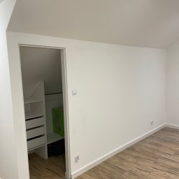 Przemysław Łabiak - Jasne wnętrze poddasza z drewnopodobną podłogą i wejściem do garderoby z białymi półkami i szufladami. Świeżo pomalowane ściany i listwy przypodłogowe.