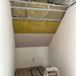 Przemysław Łabiak - Montaż sufitu podwieszanego z płyt karton-gips w skosie: widoczne profile, izolacja z wełny mineralnej i częściowo zamontowane płyty. Rusztowanie i drabina w pomieszczeniu.