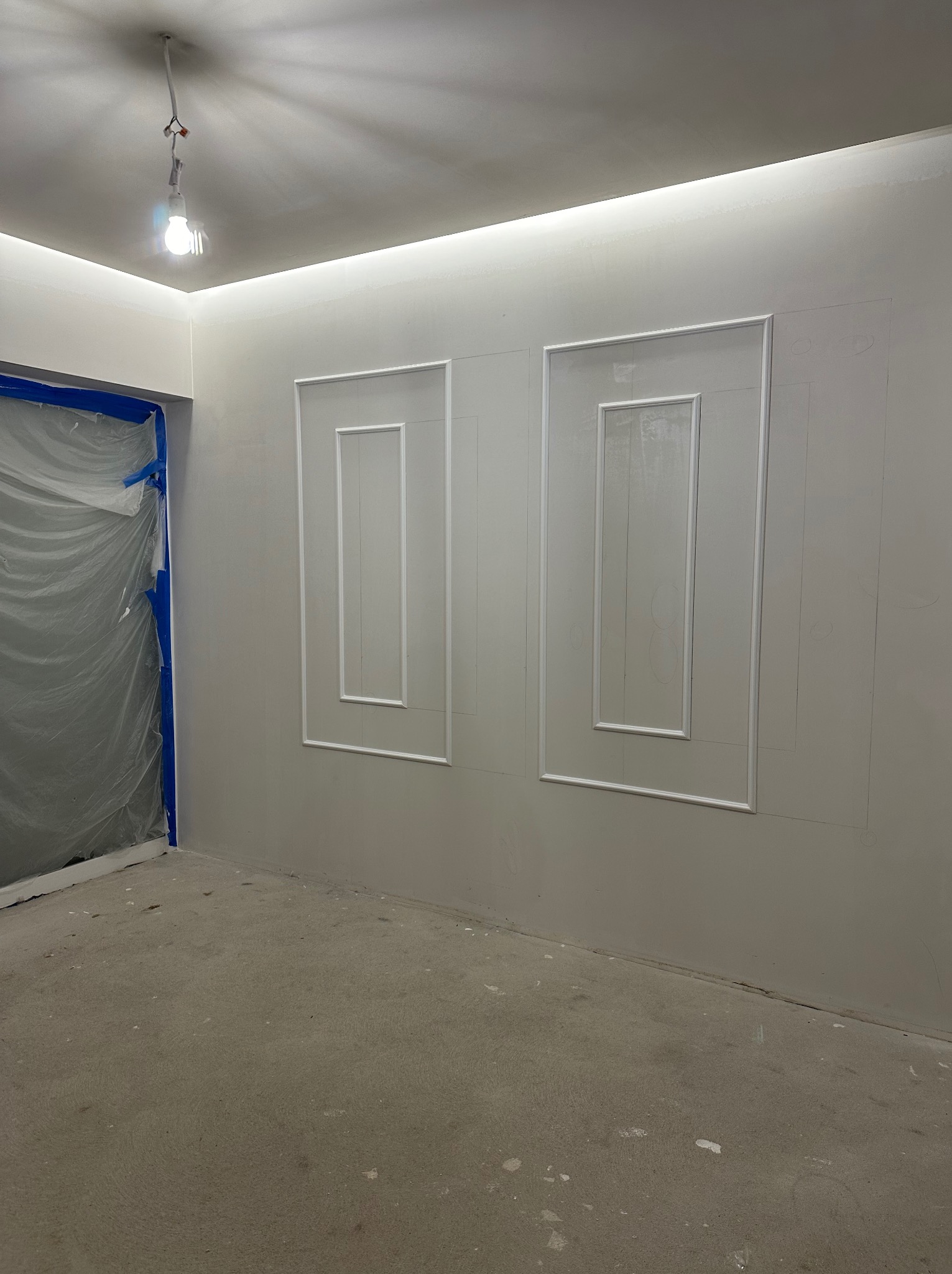 Ściana z dwoma dekoracyjnymi, białymi ramkami. Drzwi zabezpieczone folią malarską. Widoczne ślady ołówka na ścianie. Oświetlenie sufitowe LED.