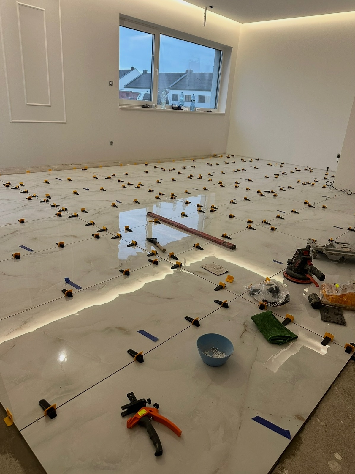 Białe, marmurowe płytki podłogowe w trakcie układania, z użyciem klipsów i poziomicy, w jasnym pomieszczeniu z oknem i listwami dekoracyjnymi.