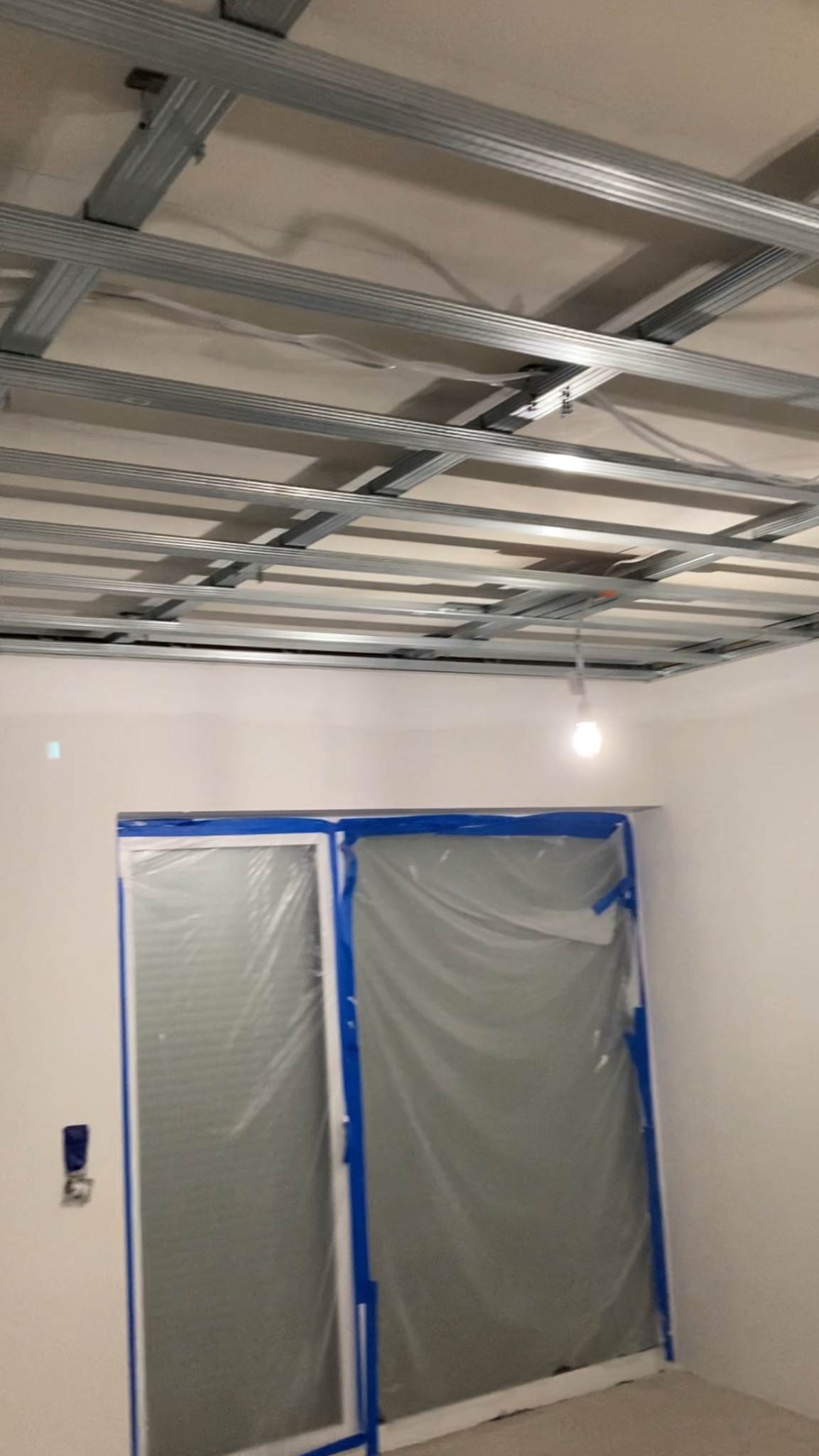 Montaż sufitu podwieszanego z płyt karton-gips. Widoczne metalowe profile konstrukcyjne. Okno zabezpieczone folią malarską. W tle biała ściana i żarówka.