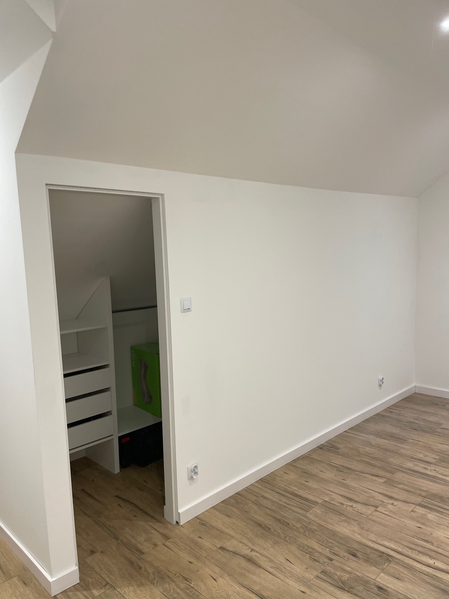 Jasne wnętrze poddasza z drewnopodobną podłogą i wejściem do garderoby z białymi półkami i szufladami. Świeżo pomalowane ściany i listwy przypodłogowe.