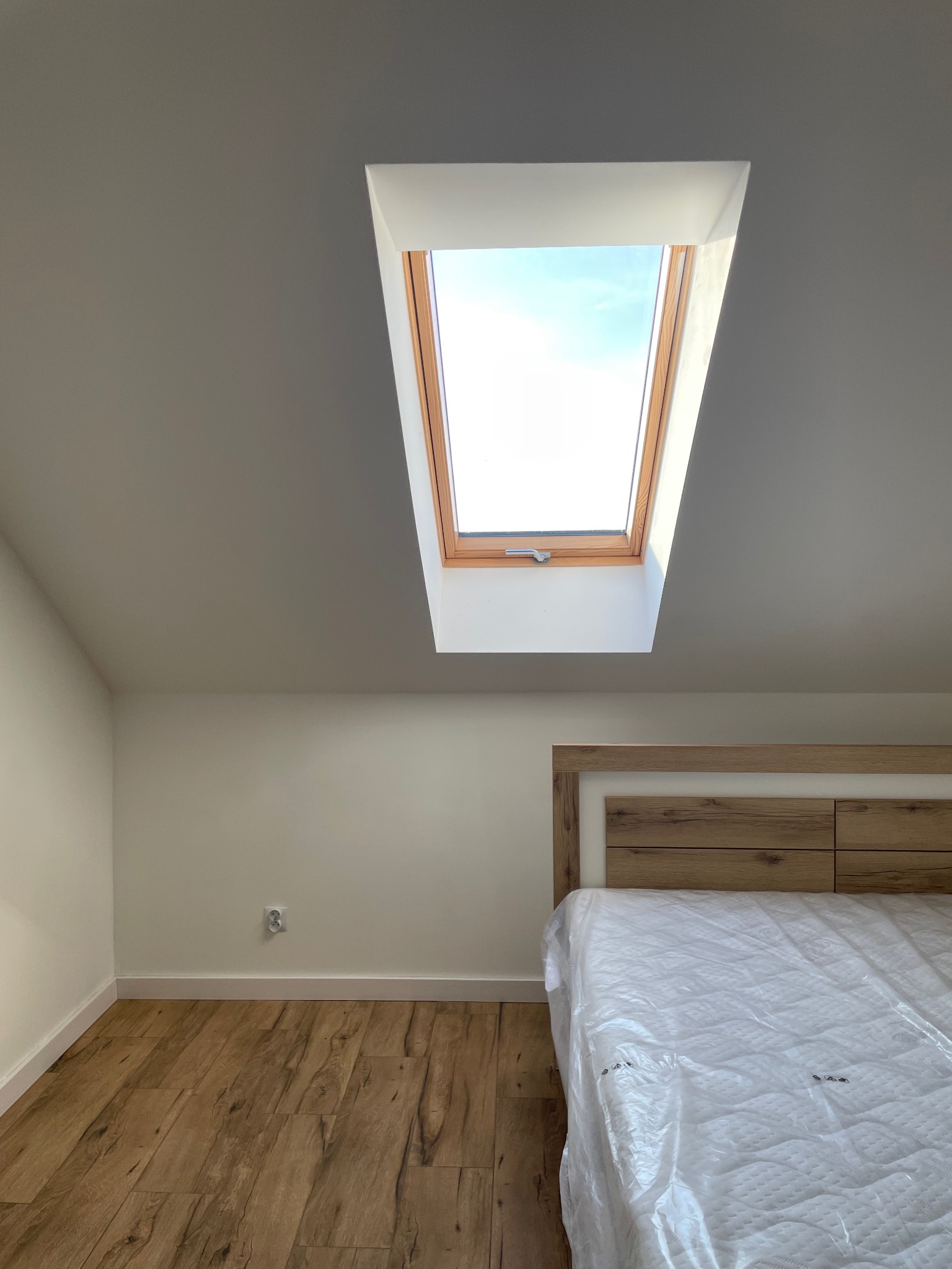 Jasne wnętrze poddasza z drewnianą podłogą i oknem dachowym. Ściany pomalowane na biało, widoczne gniazdko elektryczne. Częściowo widoczne łóżko z zagłówkiem.