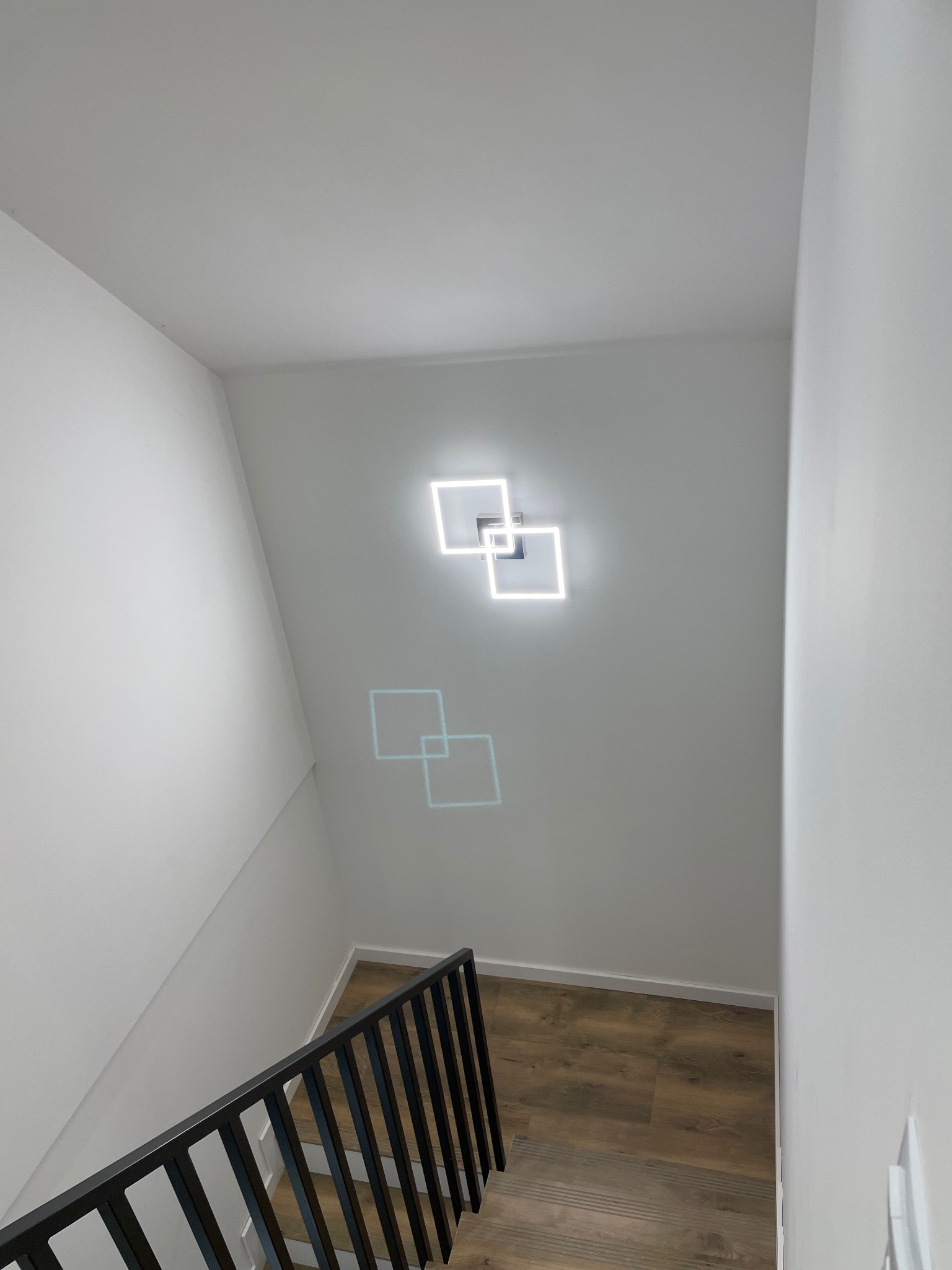 Białe ściany klatki schodowej z nowoczesnym oświetleniem LED w kształcie kwadratów, drewnopodobne panele na schodach i czarna metalowa balustrada.
