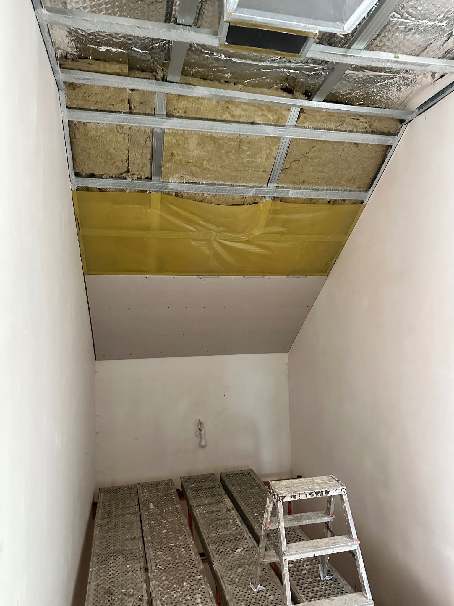 Montaż sufitu podwieszanego z płyt karton-gips w skosie: widoczne profile, izolacja z wełny mineralnej i częściowo zamontowane płyty. Rusztowanie i drabina w pomieszczeniu.