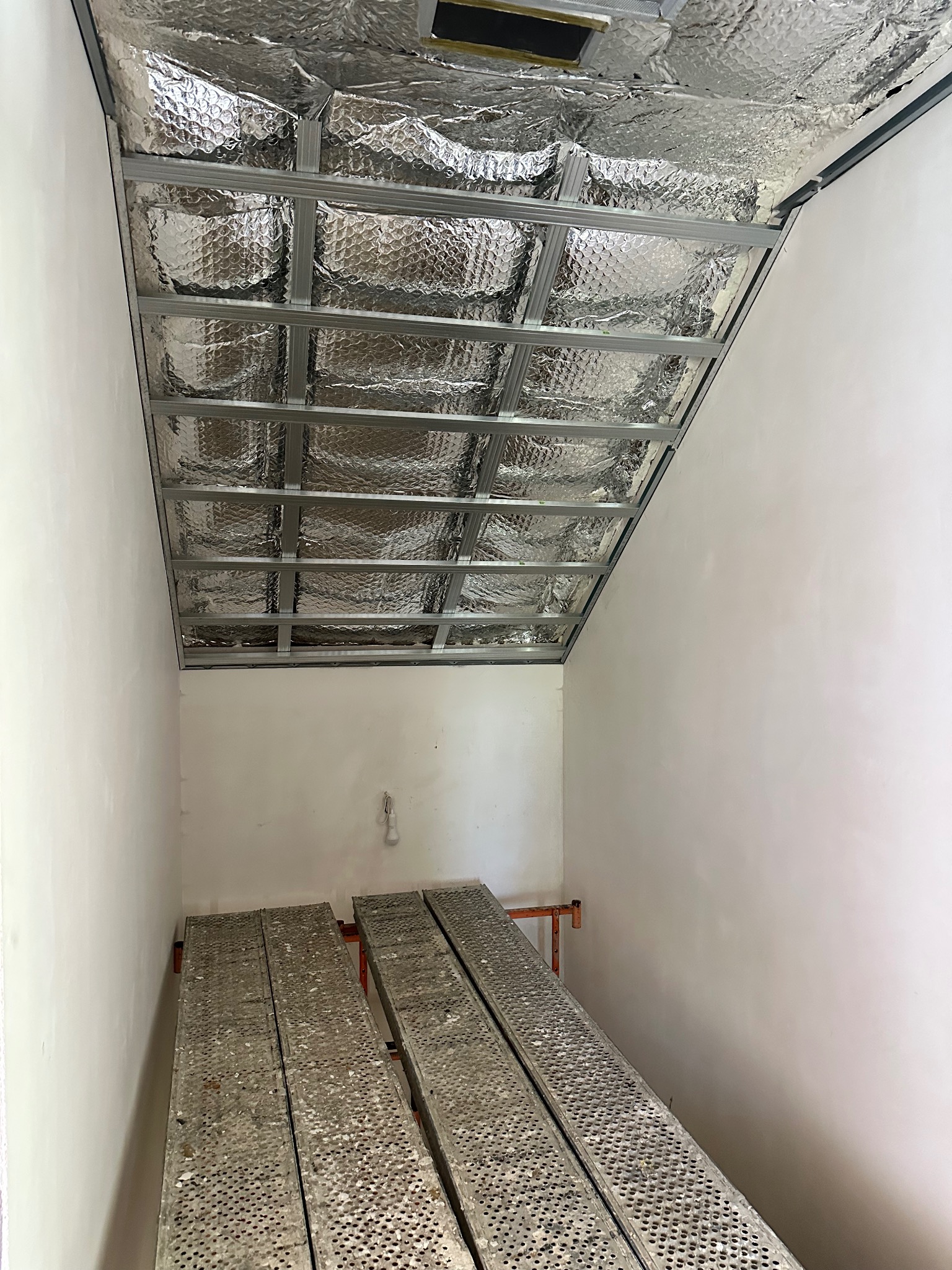 Srebrna izolacja termiczna na metalowym stelażu sufitu podwieszanego w trakcie montażu, widok z dołu na rusztowanie w pomieszczeniu z białymi ścianami.