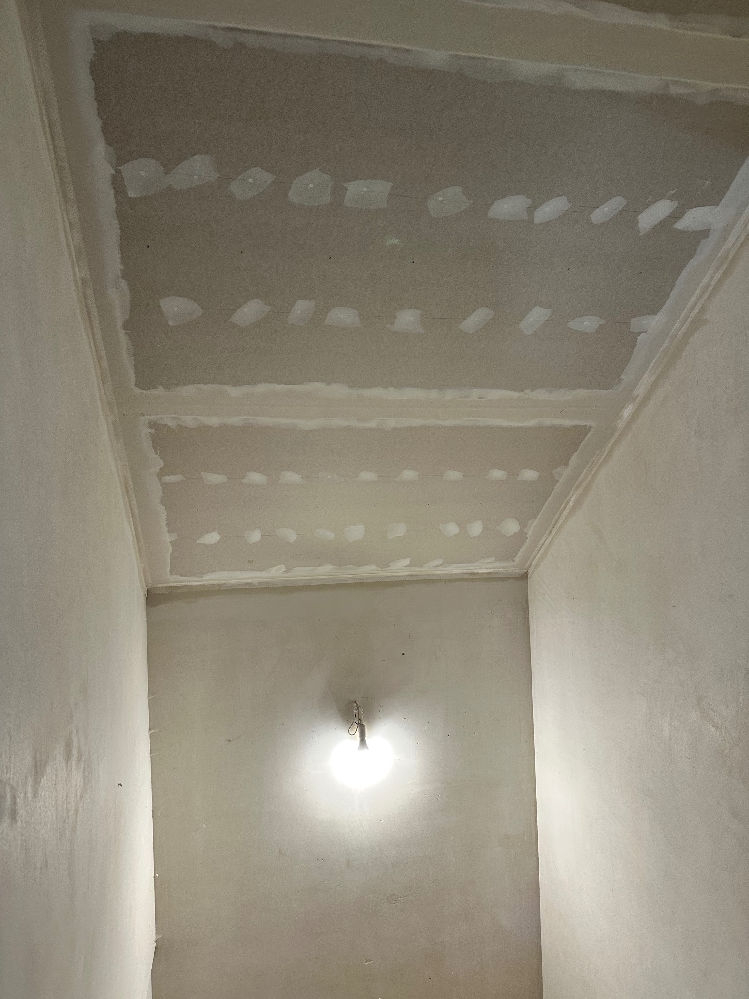 Sufit z płyt kartonowo-gipsowych, widoczne łączenia szpachlowane, przygotowany do dalszych prac wykończeniowych, oświetlony pojedynczą żarówką.
