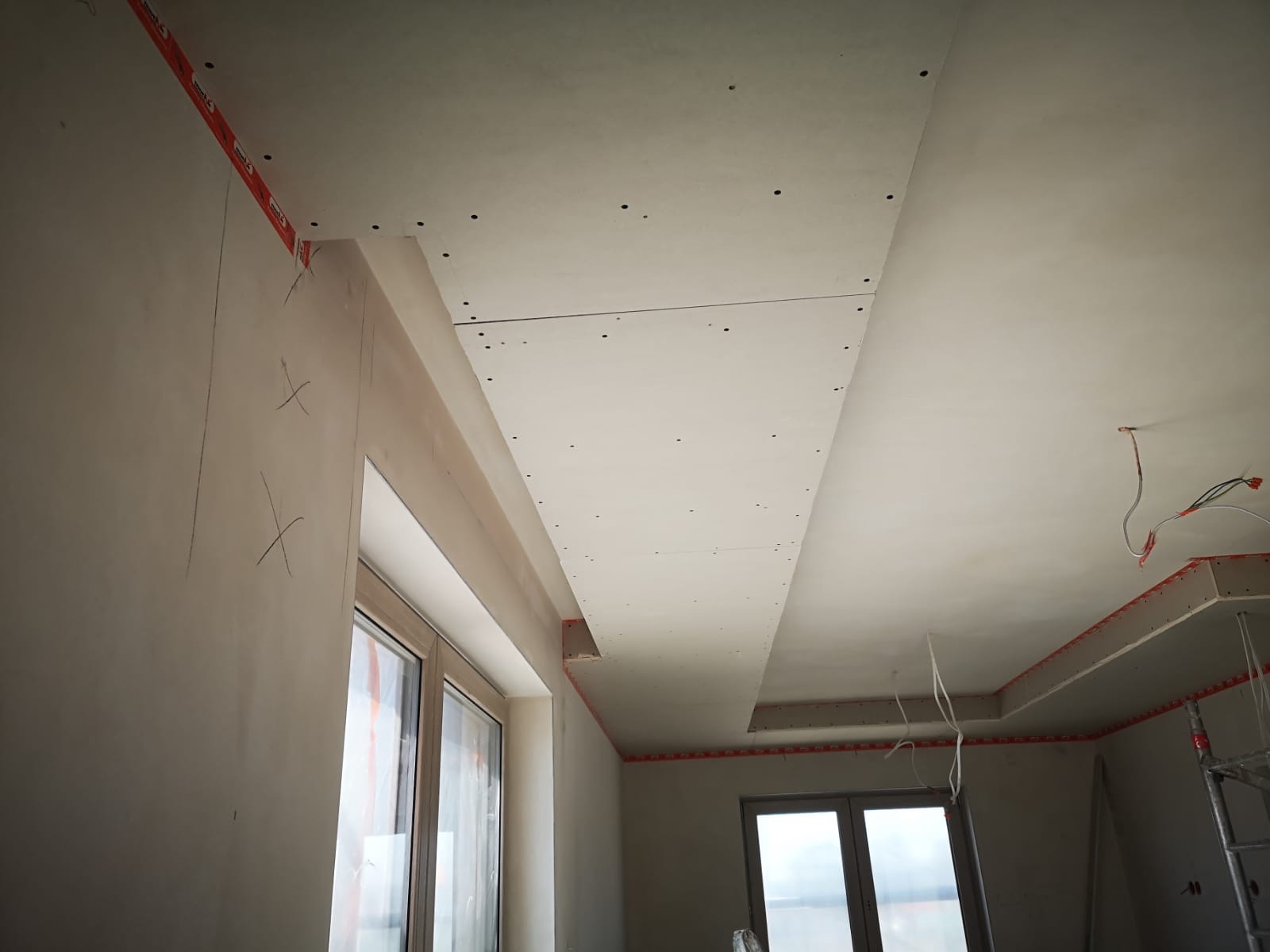 Sufit podwieszany z płyt kartonowo-gipsowych w trakcie montażu, widoczne profile i taśmy mocujące, surowe ściany z oznaczeniami, okno w tle.