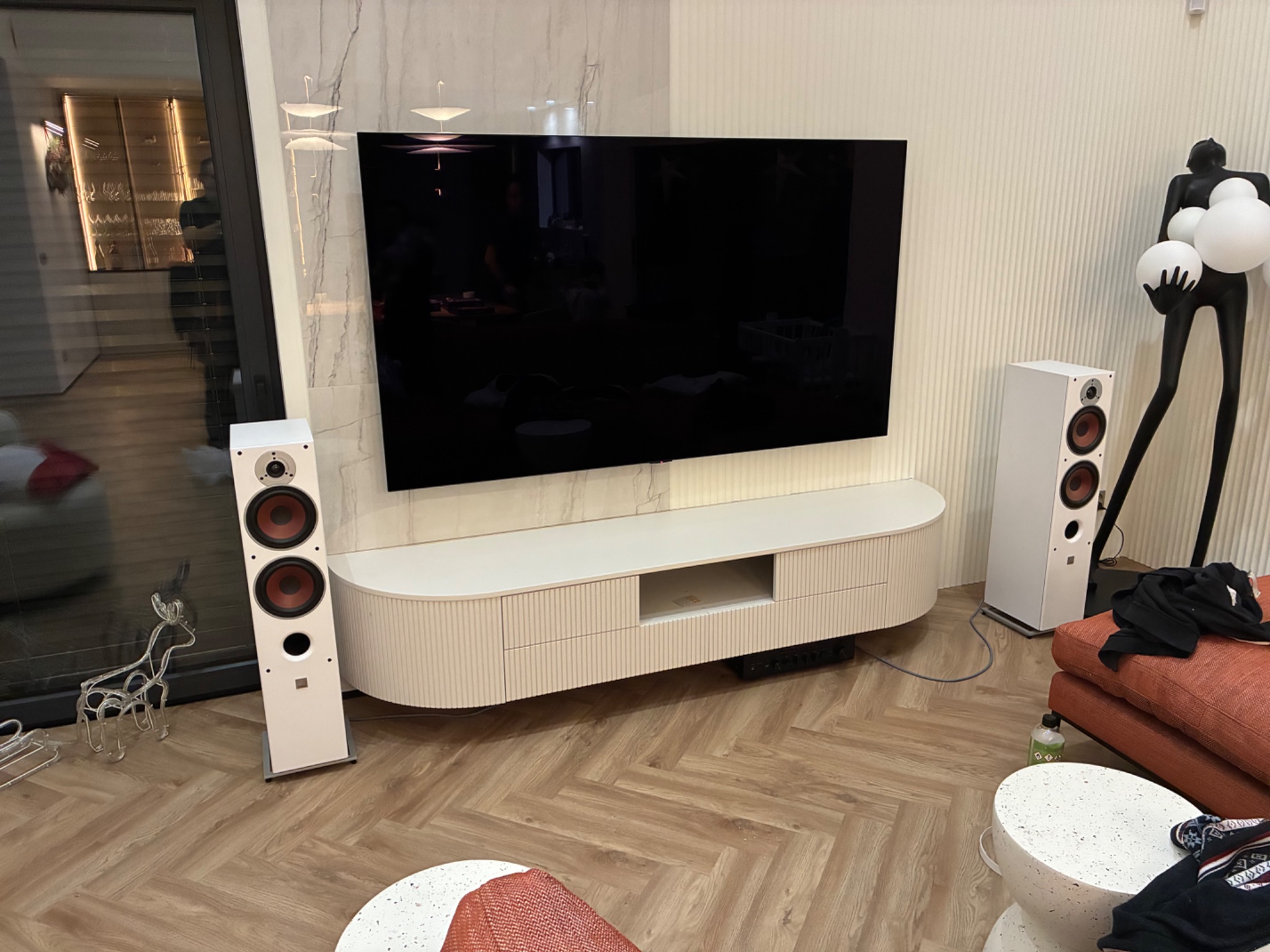 Nowoczesny salon z białymi głośnikami, szafką RTV z lamelami i marmurową ścianą. Minimalistyczny design z akcentami dekoracyjnymi.