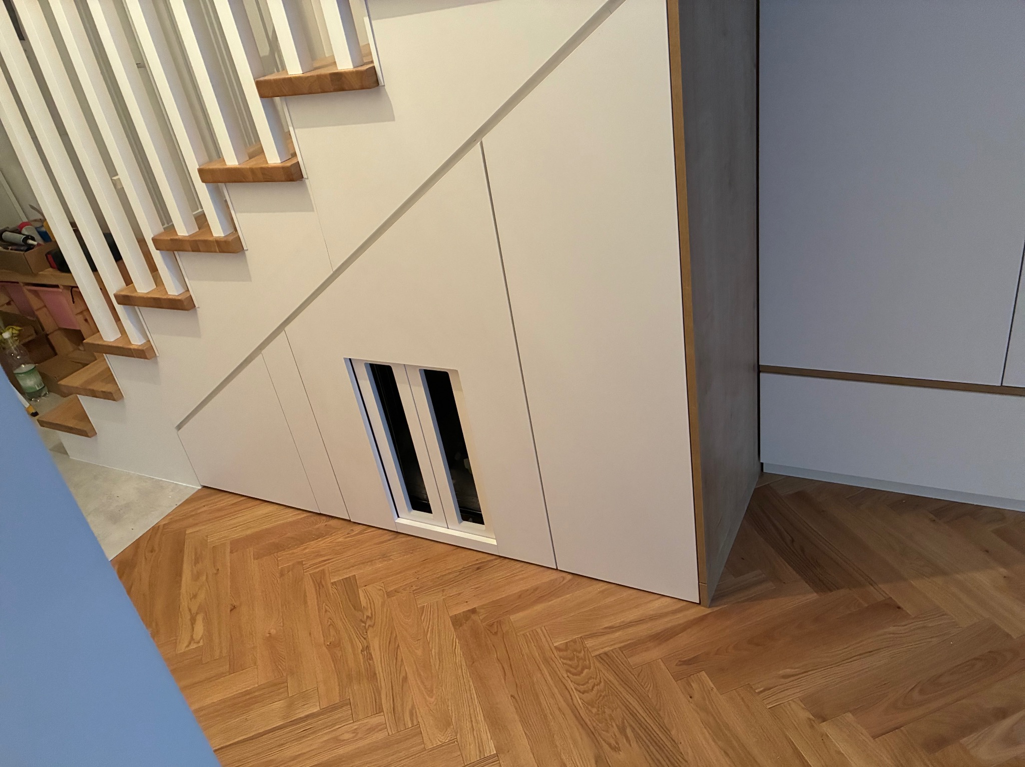 Białe schody z drewnianymi stopniami i zabudową pod schodami z małym otworem, parkiet w jodełkę na podłodze, minimalistyczny design.