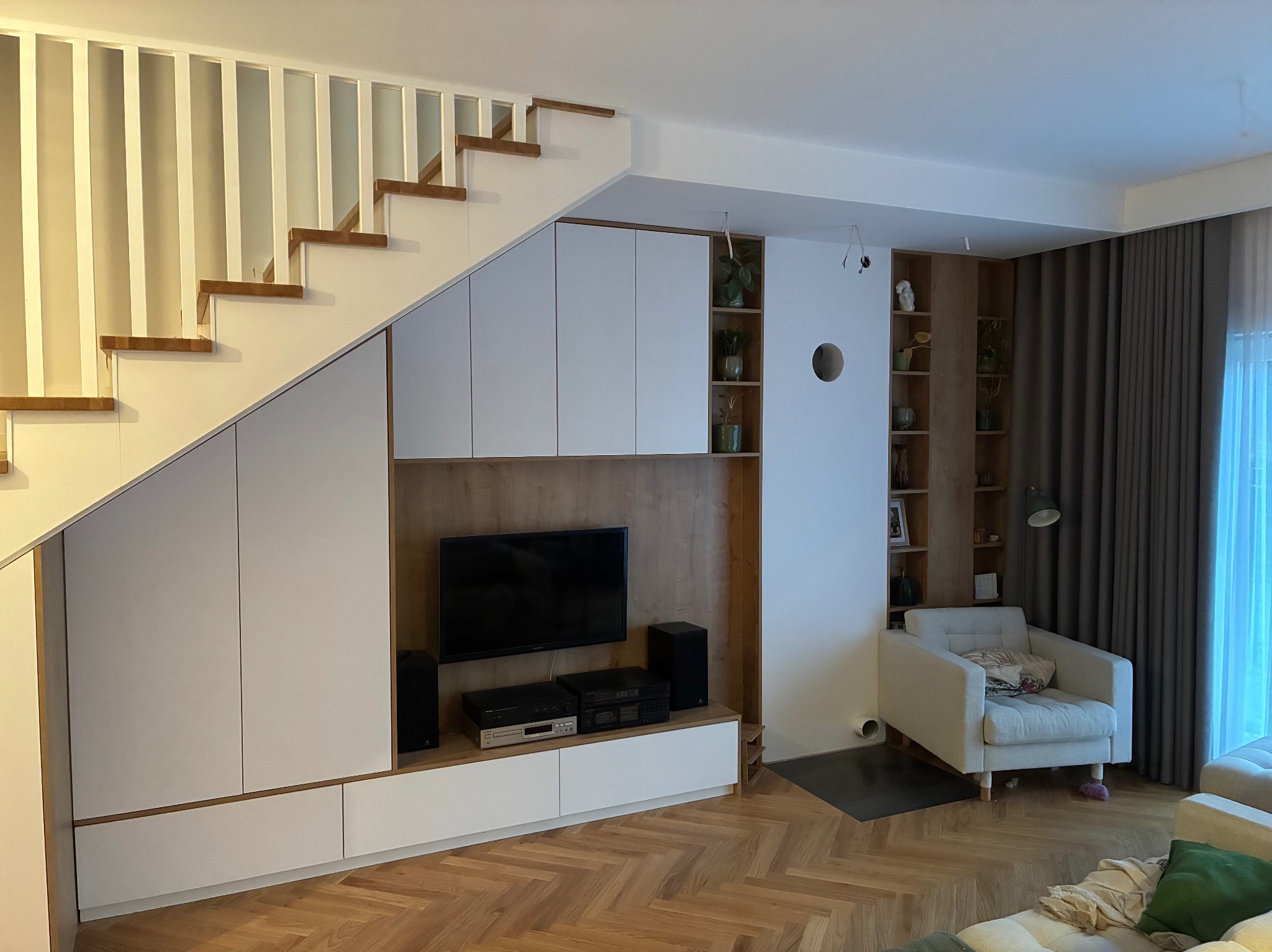 Nowoczesny salon z drewnianymi schodami i zabudową meblową pod nimi, parkiet w jodełkę, fotel i regał z roślinami. Jasne wnętrze z elementami drewna.