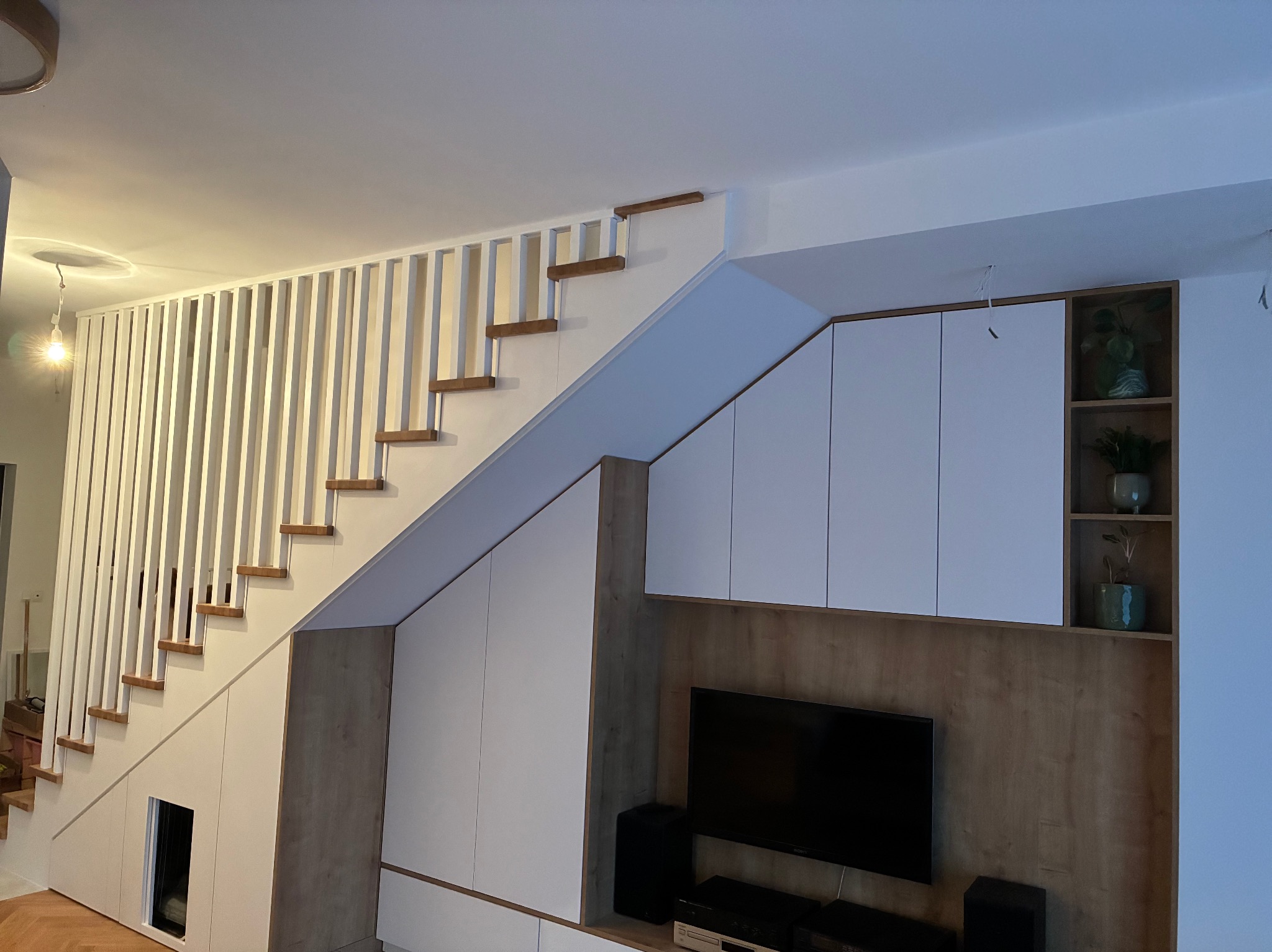 Białe schody z drewnianymi stopniami i zabudową pod schodami, wkomponowane w nowoczesne wnętrze z szafkami i półkami na rośliny.
