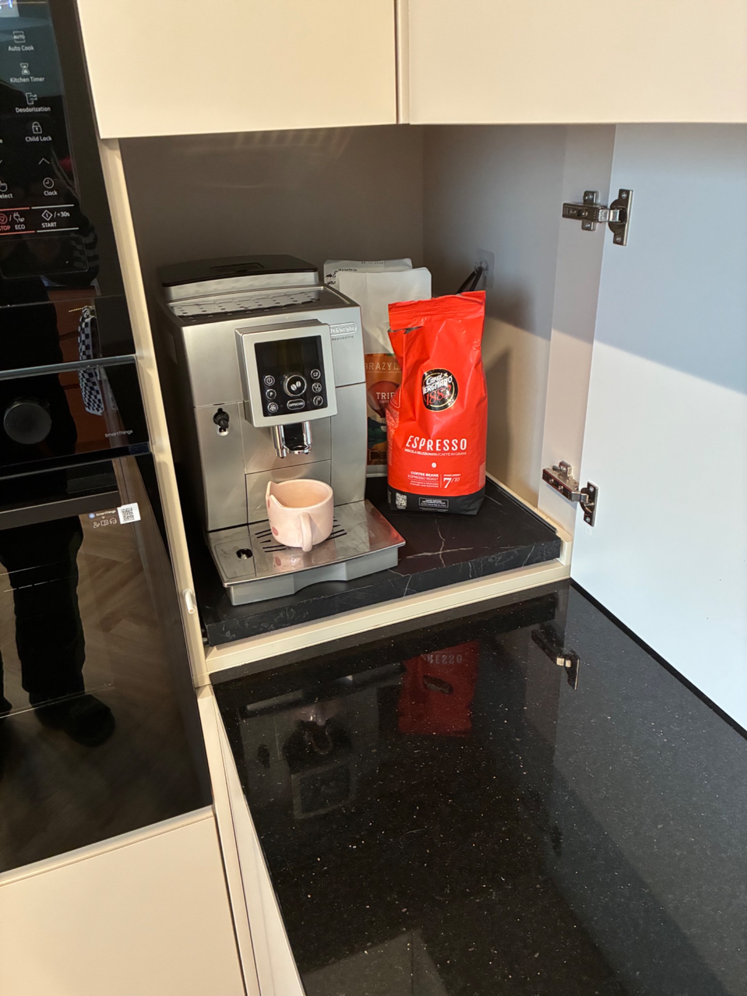 Wnęka kuchenna z ekspresem do kawy DeLonghi, paczką kawy Vergnano Espresso i otwartymi białymi drzwiczkami szafki, na blacie z czarnego, błyszczącego kamienia.