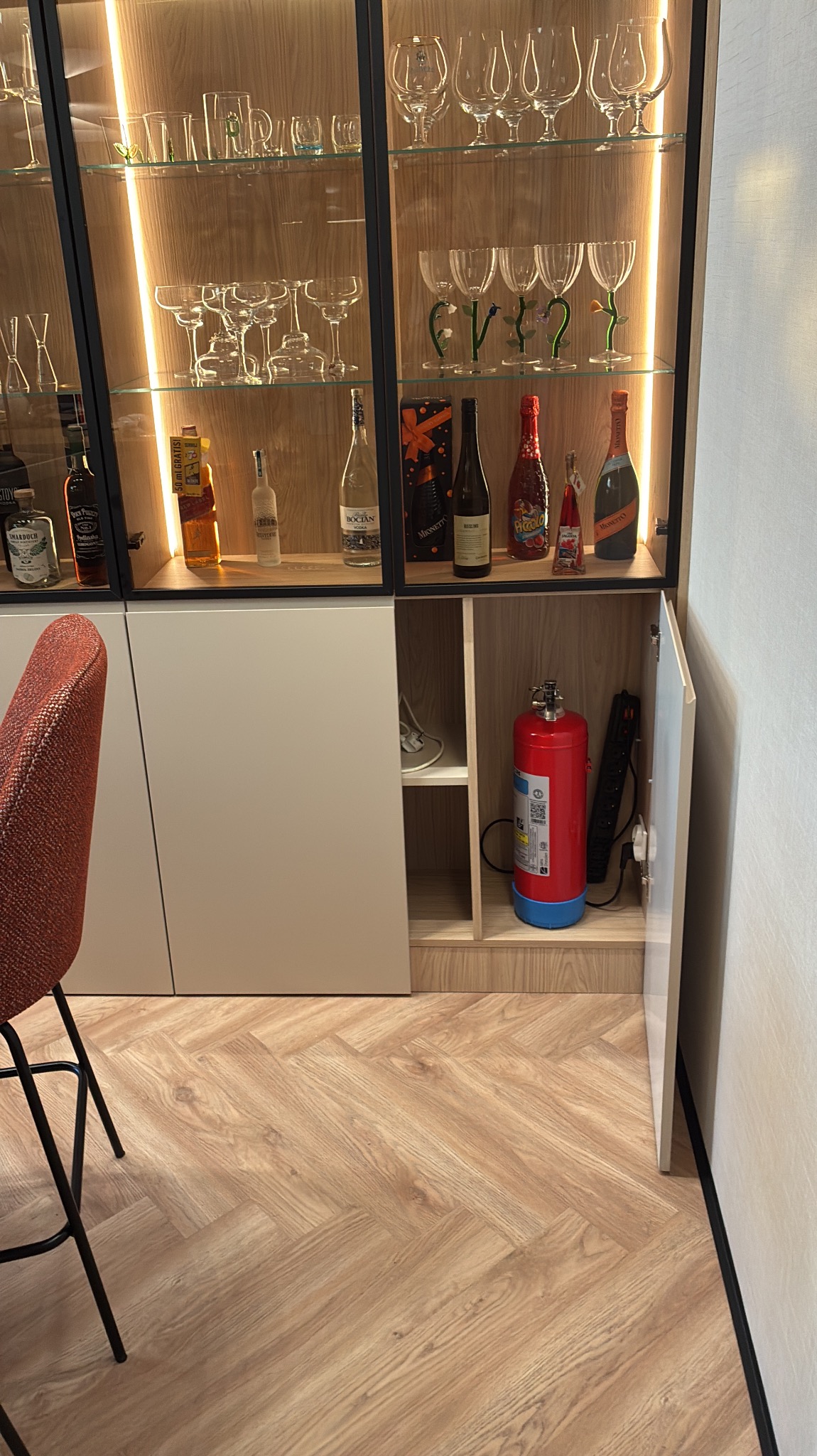 Nowoczesna szafa z przeszklonymi witrynami, podświetlonymi półkami, eksponująca szkło i alkohole. W dolnej części szafki gaśnica i listwa zasilająca. Parkiet w jodełkę.