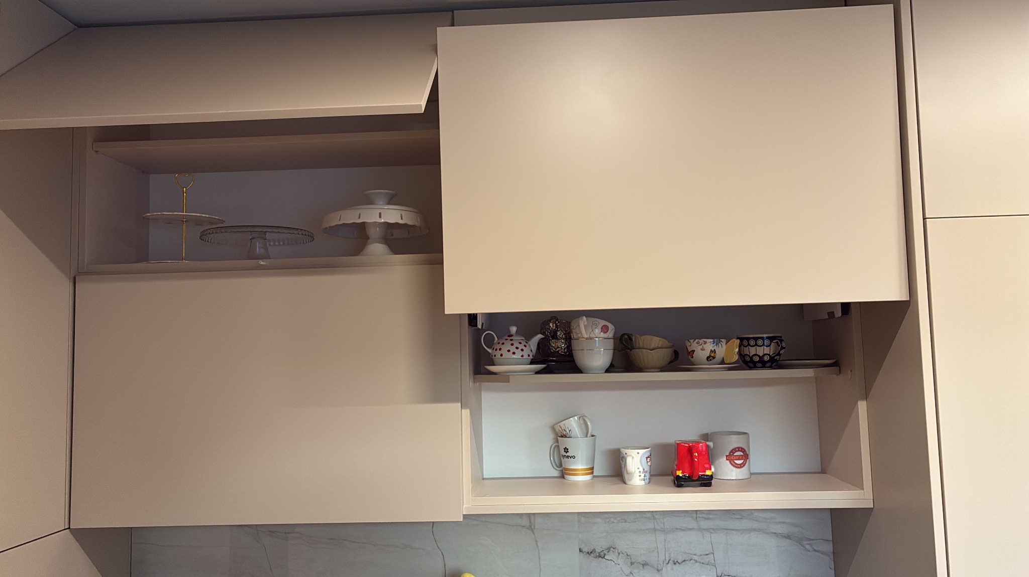 Beżowe szafki kuchenne z otwartymi drzwiczkami, ukazujące półki z naczyniami i dekoracjami. Minimalistyczny design, jasne wnętrze, marmurowy backsplash.
