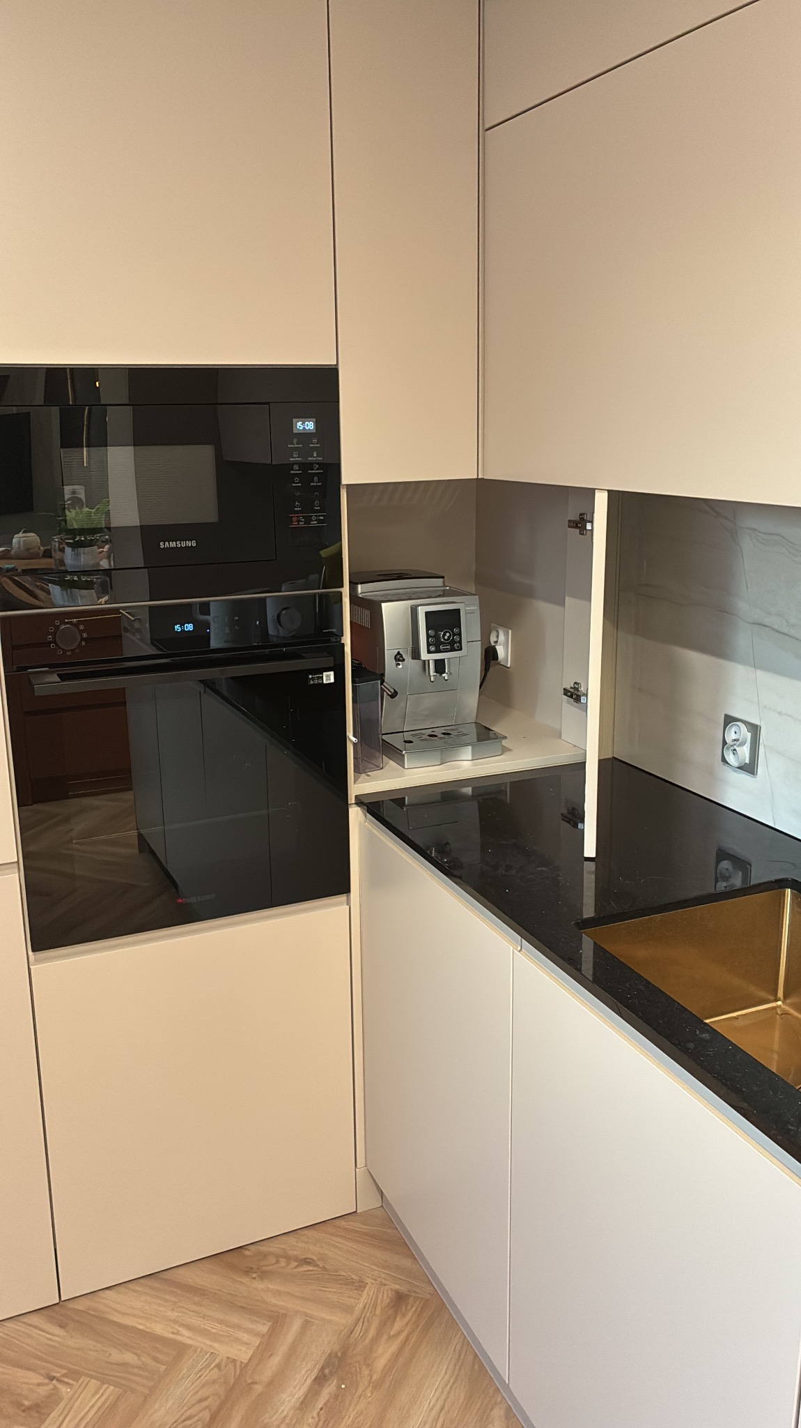 Jasna zabudowa kuchenna z czarnym blatem, wnęką na ekspres do kawy i piekarnikiem Samsung. Parkiet ułożony w jodełkę na podłodze.