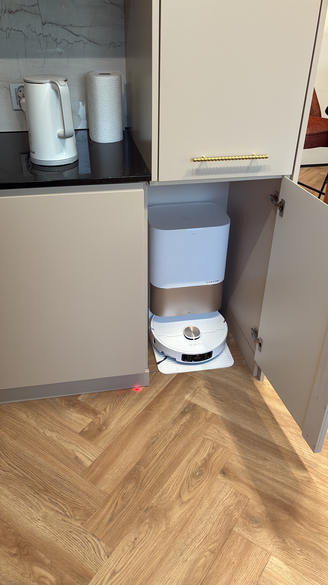 Biała szafka kuchenna z otwartymi drzwiami, wewnątrz stacja dokująca robota sprzątającego Dreame. Na blacie czajnik i ręcznik papierowy.