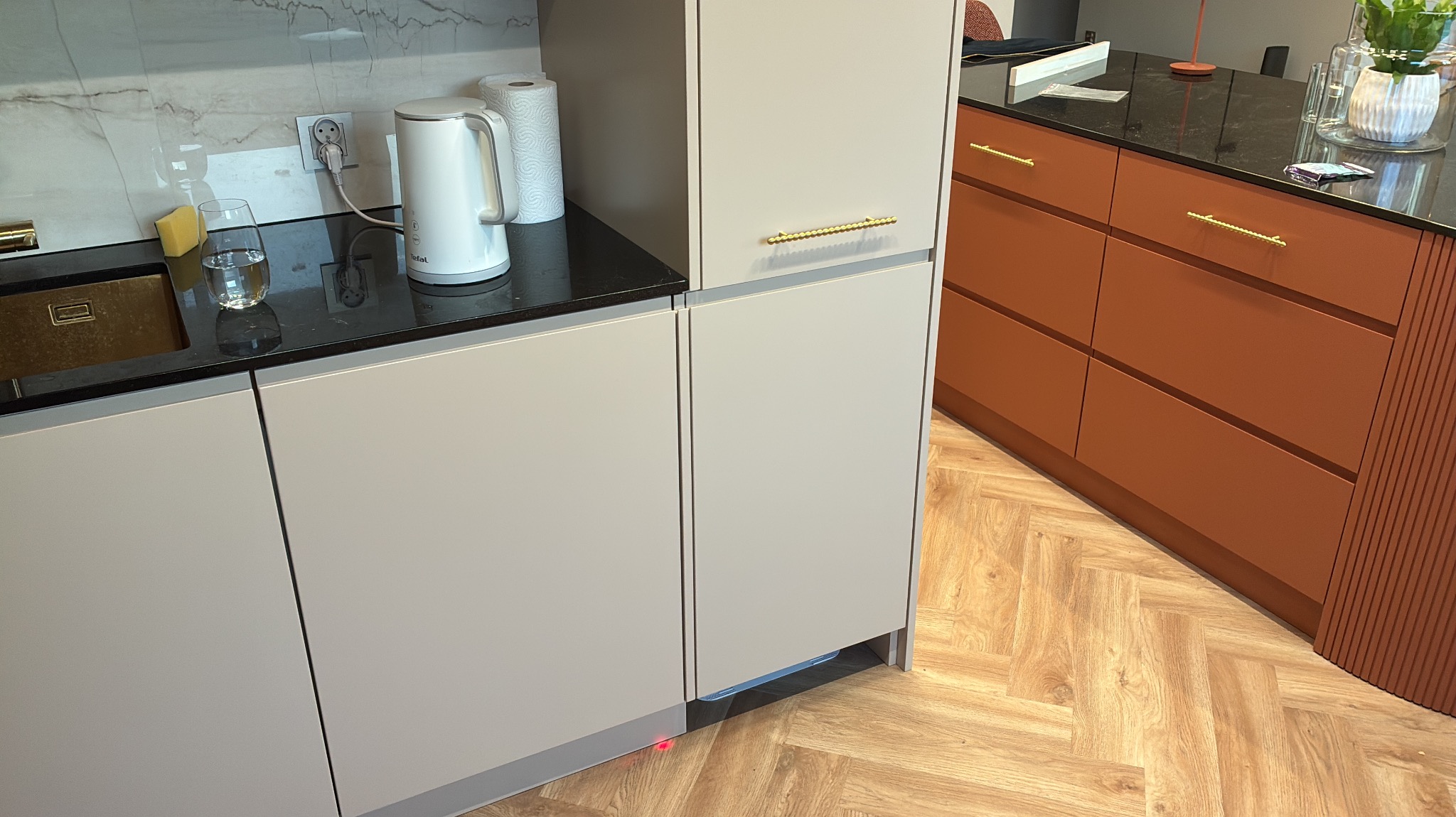 Nowoczesna kuchnia z jasnymi szafkami i czarnym blatem, uzupełniona o złote uchwyty. Widoczny czajnik i elementy wyposażenia. Parkiet w jodełkę na podłodze.