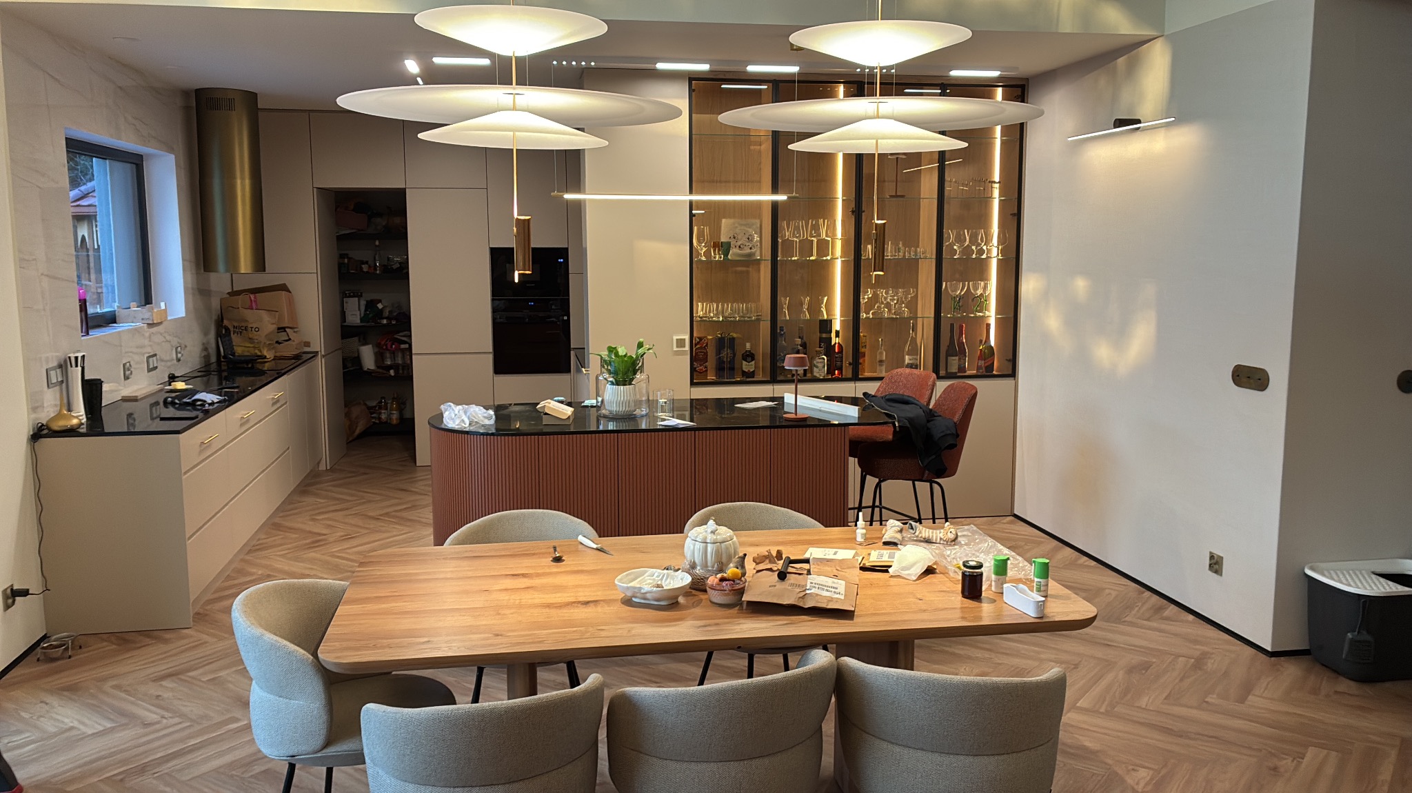 Nowoczesna kuchnia z wyspą i jadalnią, oświetlona lampami w kształcie kapeluszy. Parkiet w jodełkę, beżowe szafki i przeszklona witryna z podświetleniem.